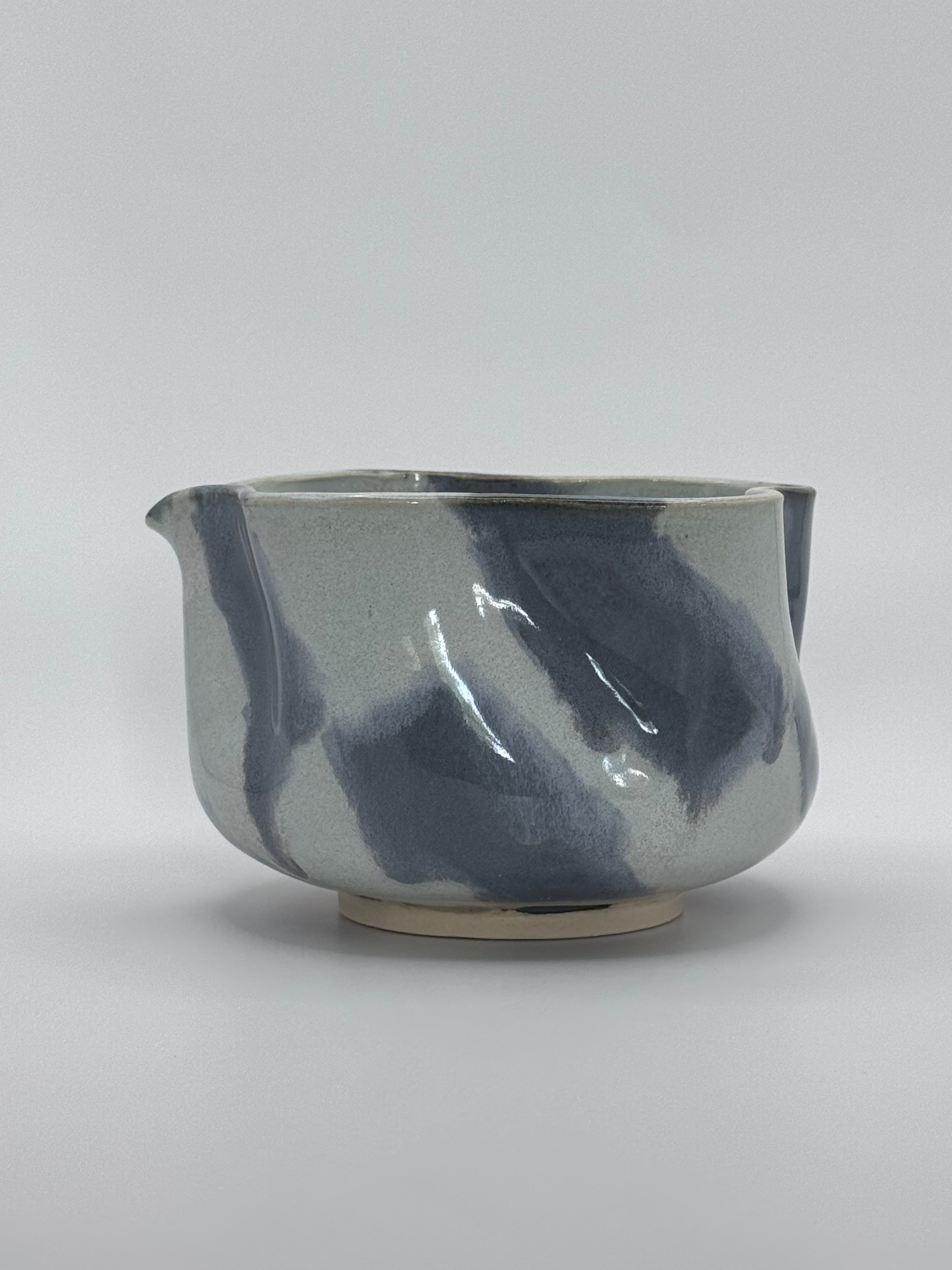 A handmade ceramic bowl with a wavy rim and a small footed base, featuring a glossy glaze with abstract slated blue and light blue brushstrokes over a white clay base, set against a plain light gray background.