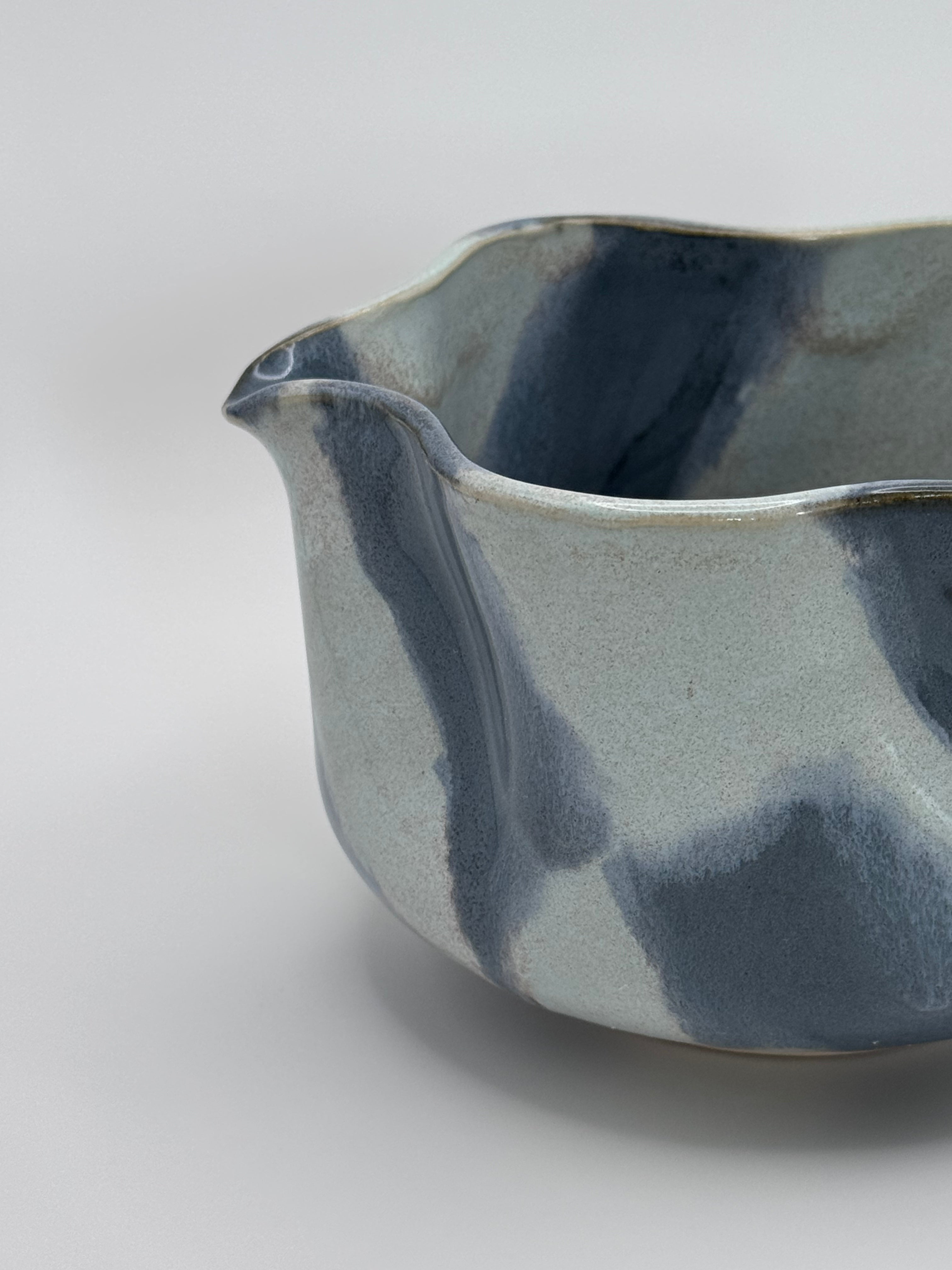 A close-up of A handmade ceramic bowl with a wavy rim and a small footed base, featuring a glossy glaze with abstract slated blue and light blue brushstrokes over a white clay base, set against a plain light gray background.