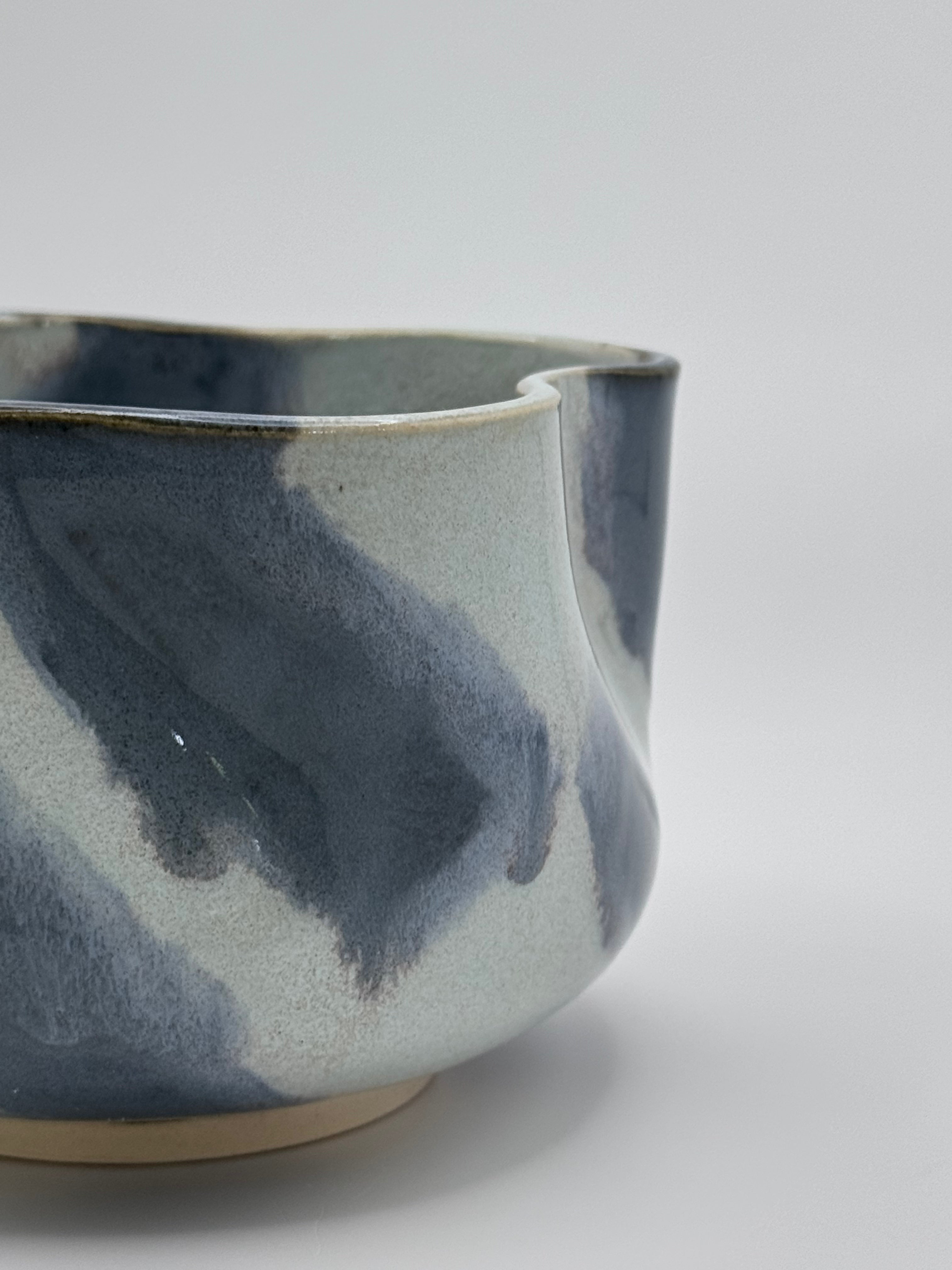 A close-up of A handmade ceramic bowl with a wavy rim and a small footed base, featuring a glossy glaze with abstract slated blue and light blue brushstrokes over a white clay base, set against a plain light gray background.