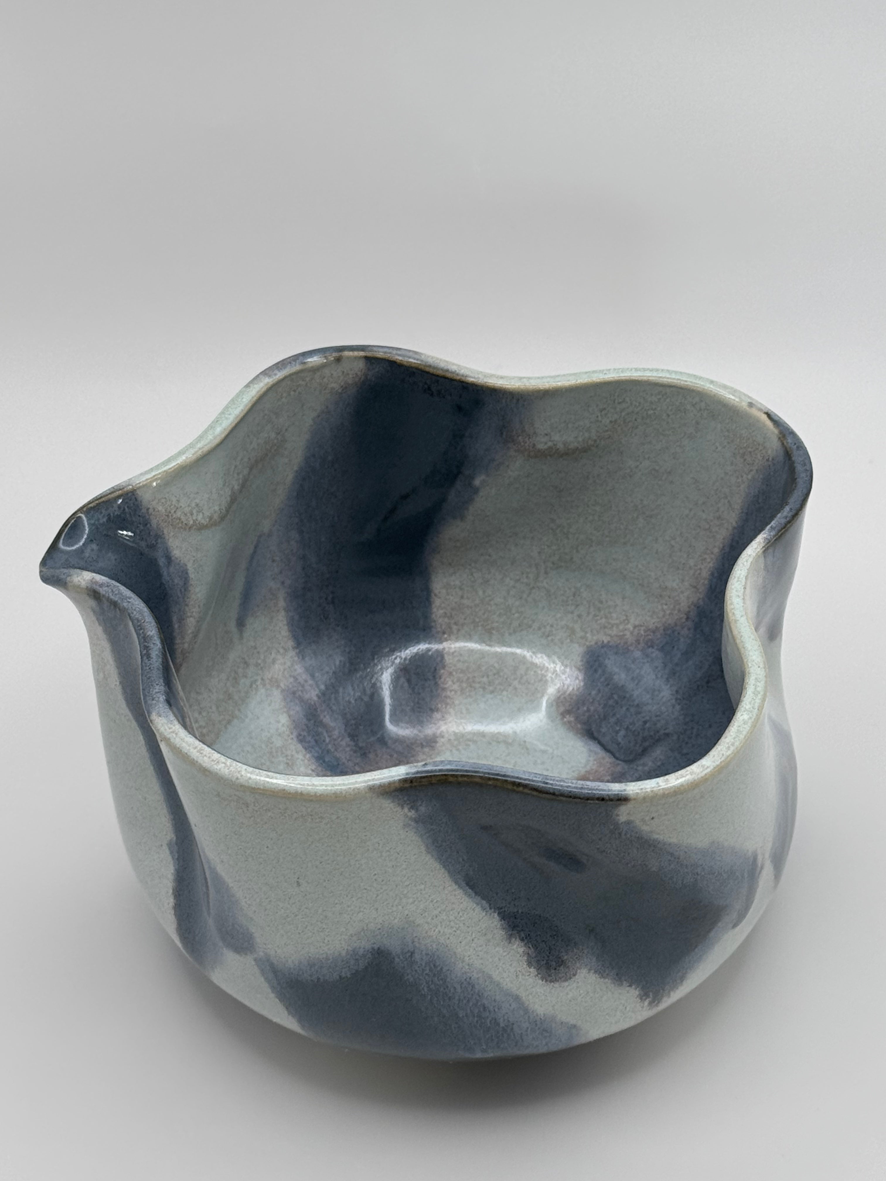 A handmade ceramic bowl with a wavy rim and a small footed base, featuring a glossy glaze with abstract slated blue and light blue brushstrokes over a white clay base, set against a plain light gray background.