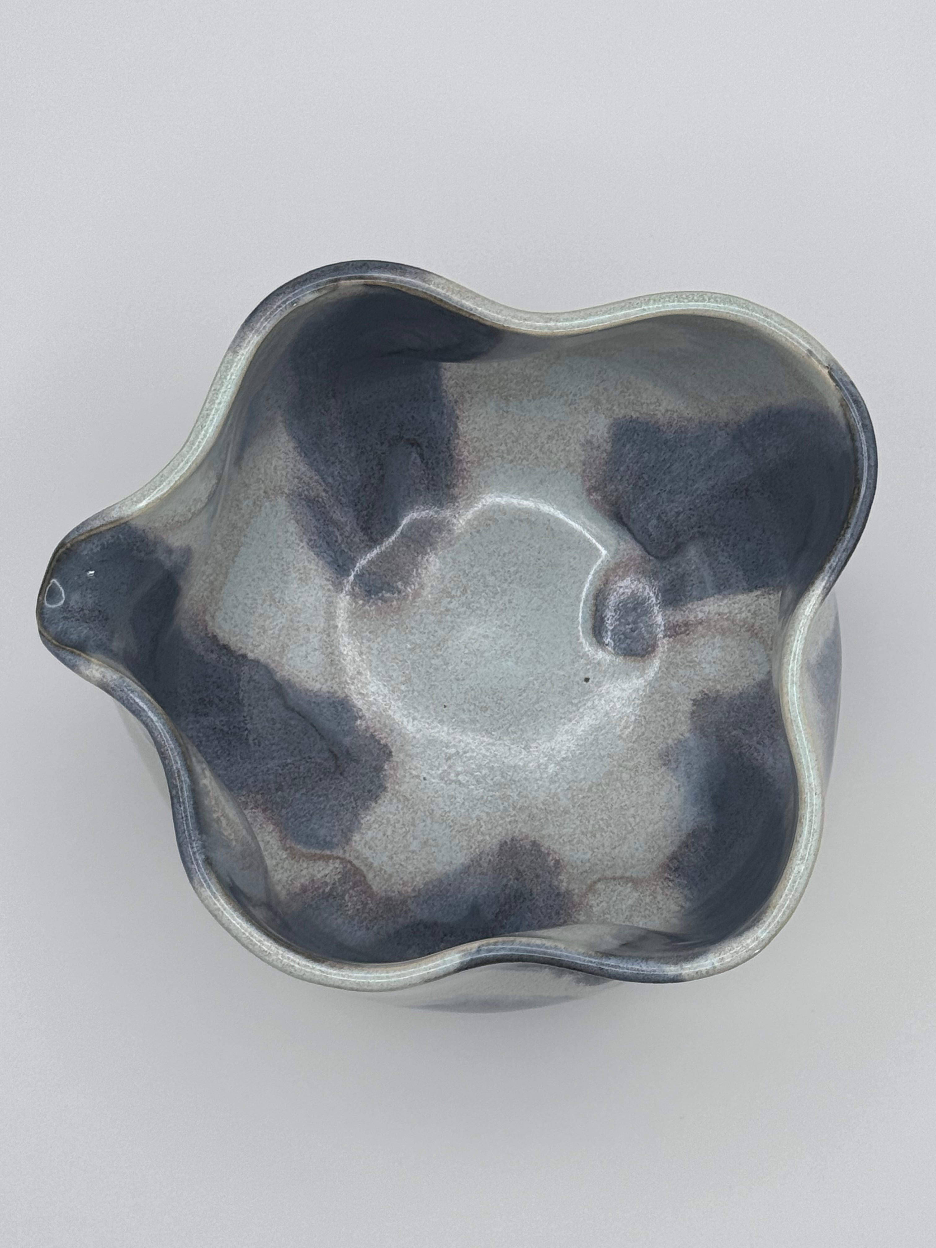 Top view of A handmade ceramic bowl with a wavy rim and a small footed base, featuring a glossy glaze with abstract slated blue and light blue brushstrokes over a white clay base and dusty rose specks on the interior, set against a plain light gray background.