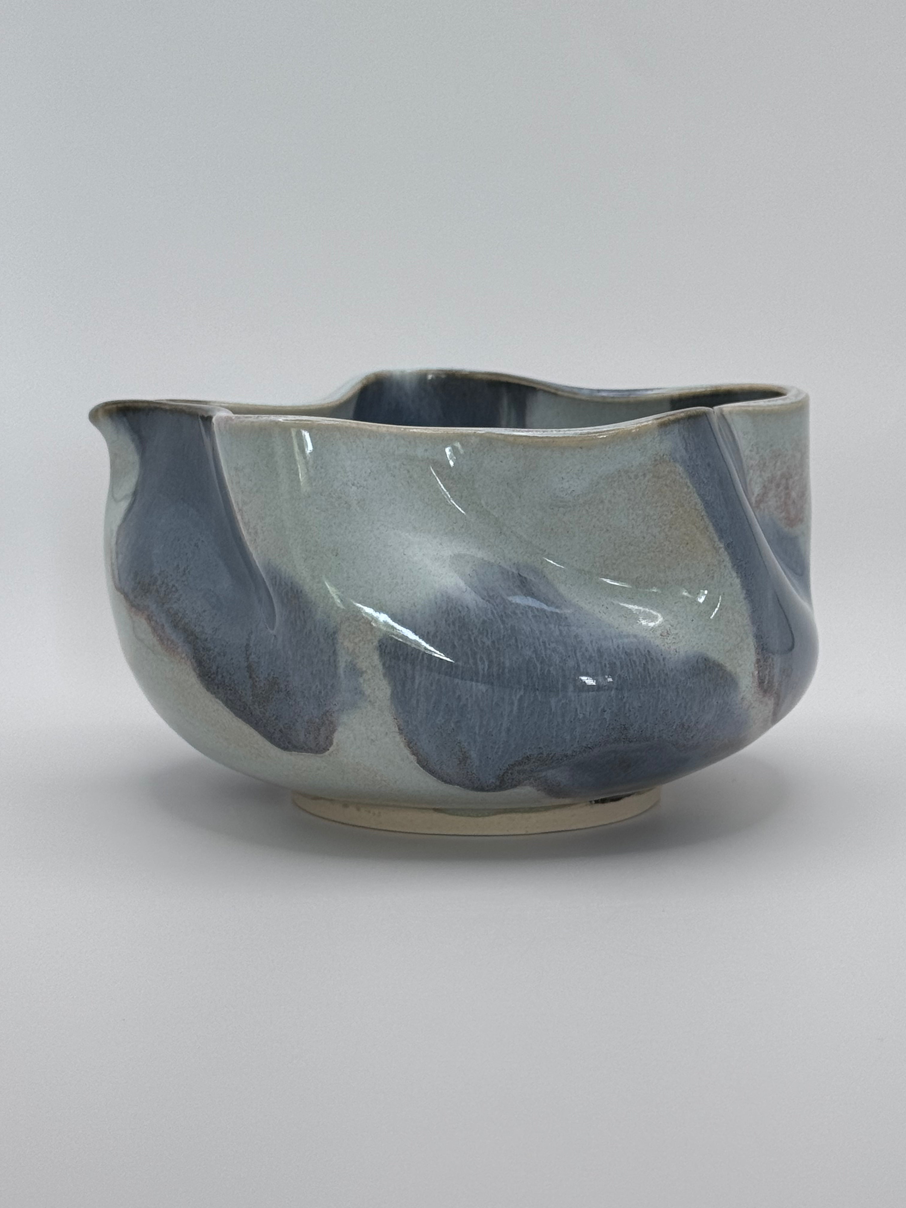 A handmade ceramic bowl with a wavy rim and a small footed base, featuring a glossy glaze with abstract slated blue and light blue brushstrokes over a white clay base, set against a plain light gray background.