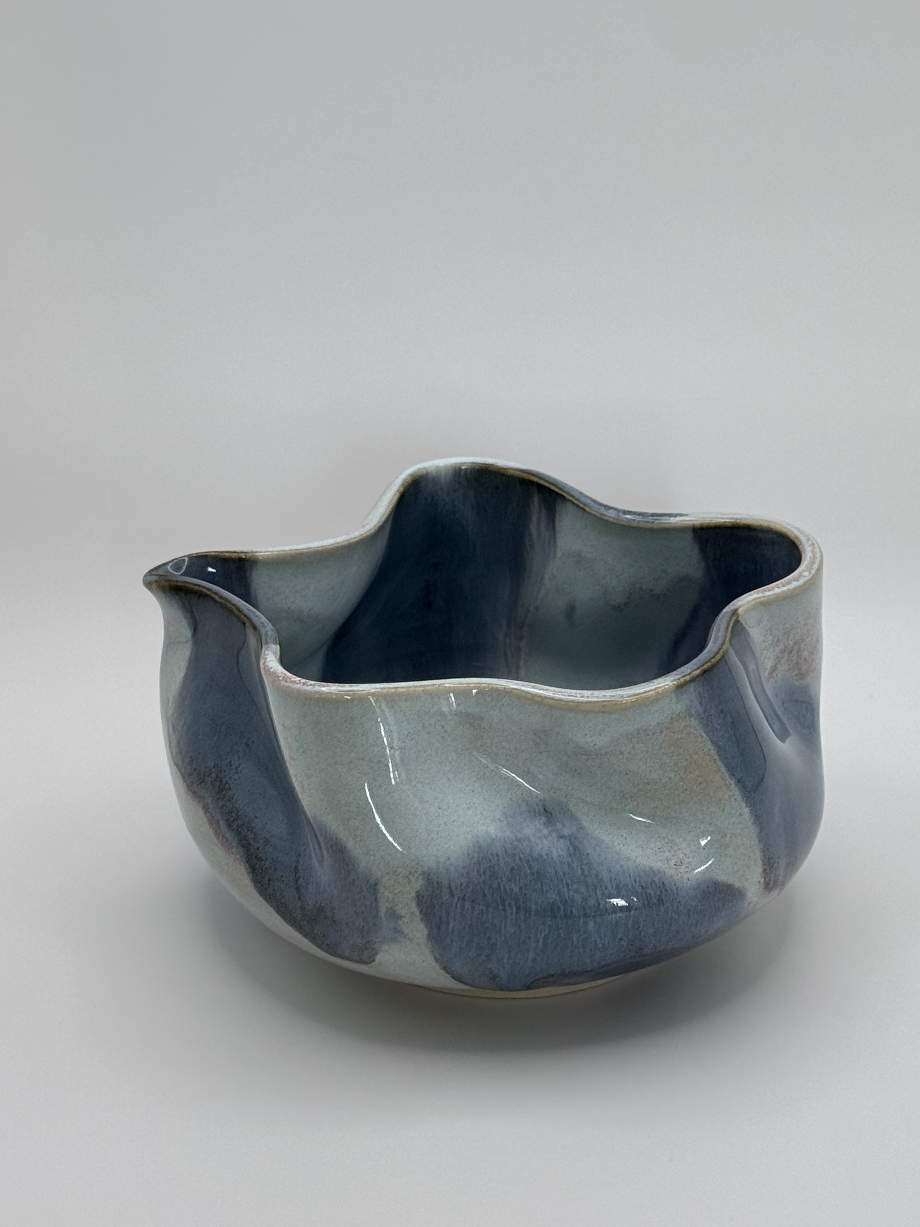 A handmade ceramic bowl with a wavy rim and a small footed base, featuring a glossy glaze with abstract slated blue and light blue brushstrokes over a white clay base, set against a plain light gray background.