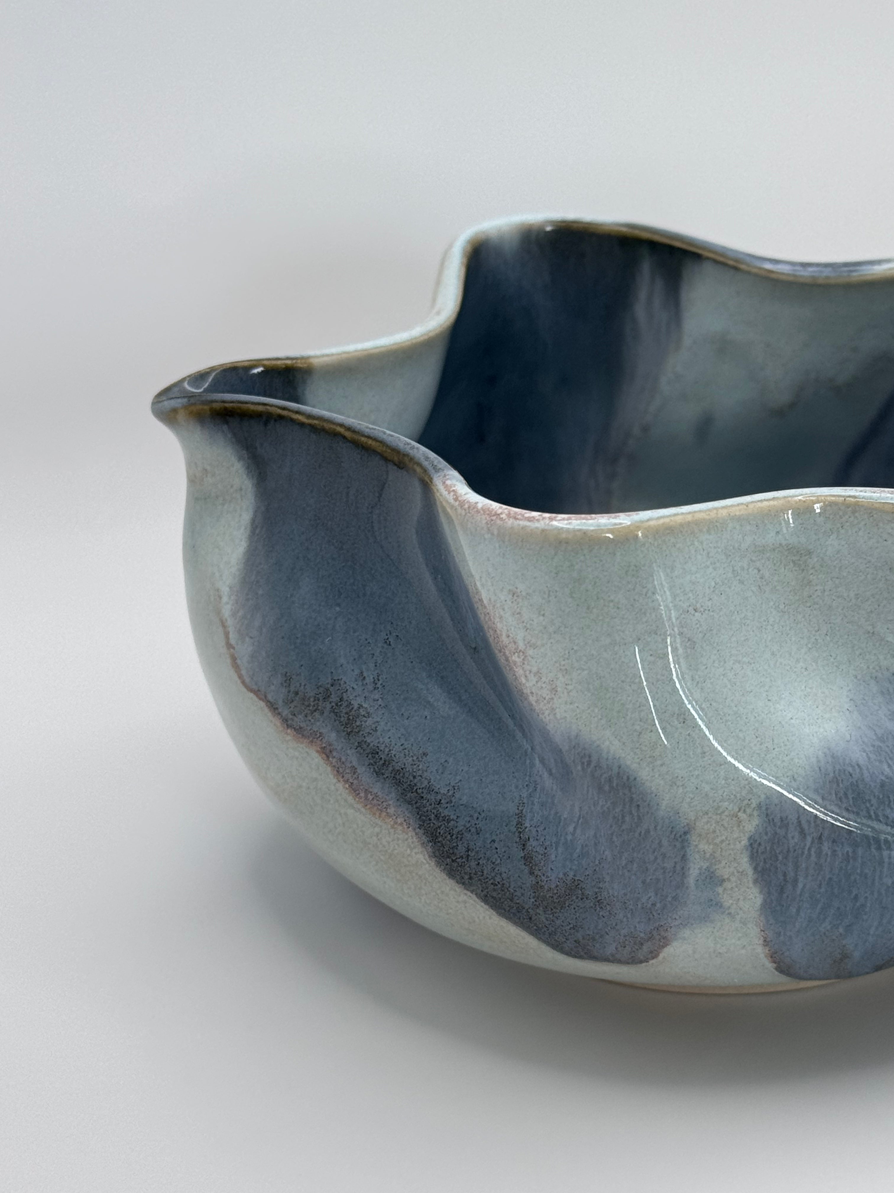A close-up of A handmade ceramic bowl with a wavy rim and a small footed base, featuring a glossy glaze with abstract slated blue and light blue brushstrokes over a white clay base, set against a plain light gray background.