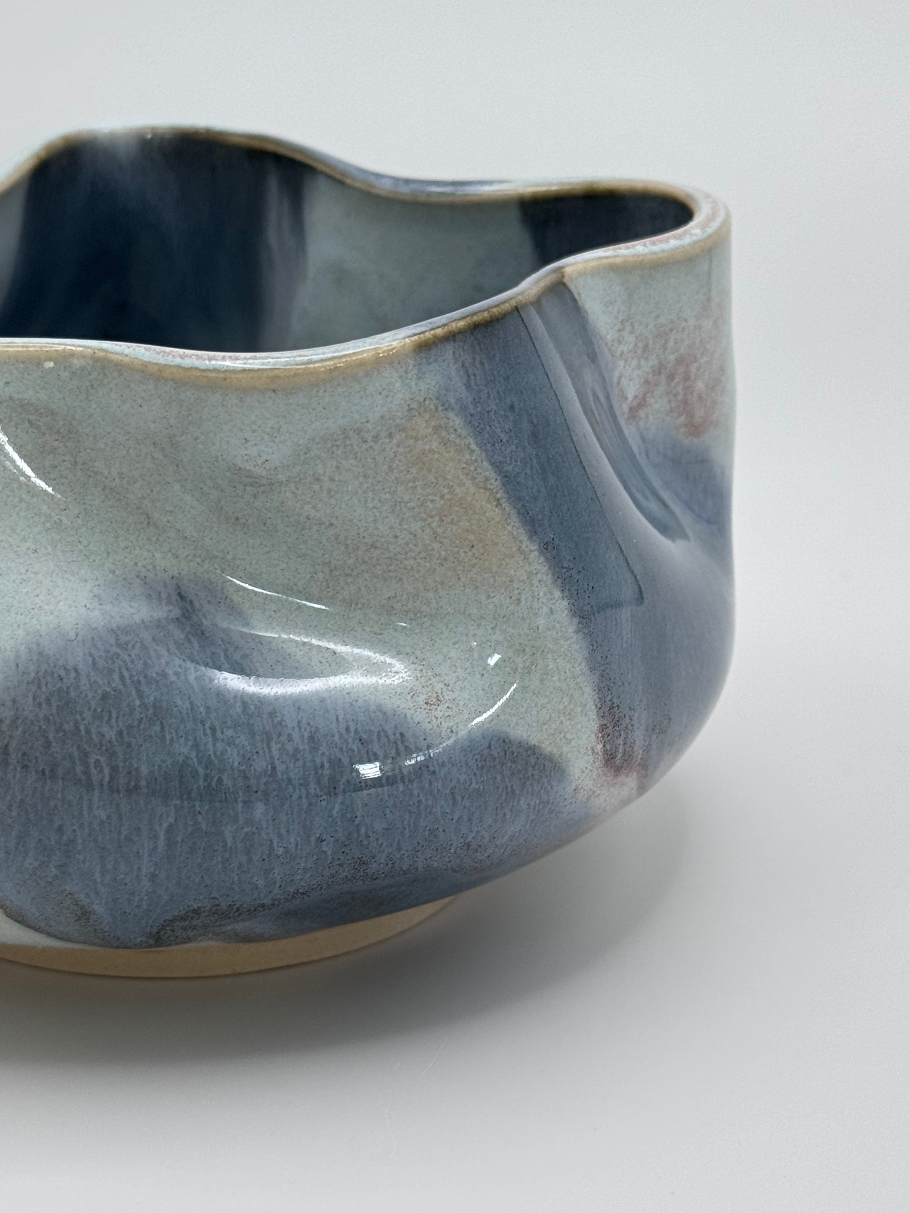 A close-up of A handmade ceramic bowl with a wavy rim and a small footed base, featuring a glossy glaze with abstract slated blue and light blue brushstrokes over a white clay base, set against a plain light gray background.