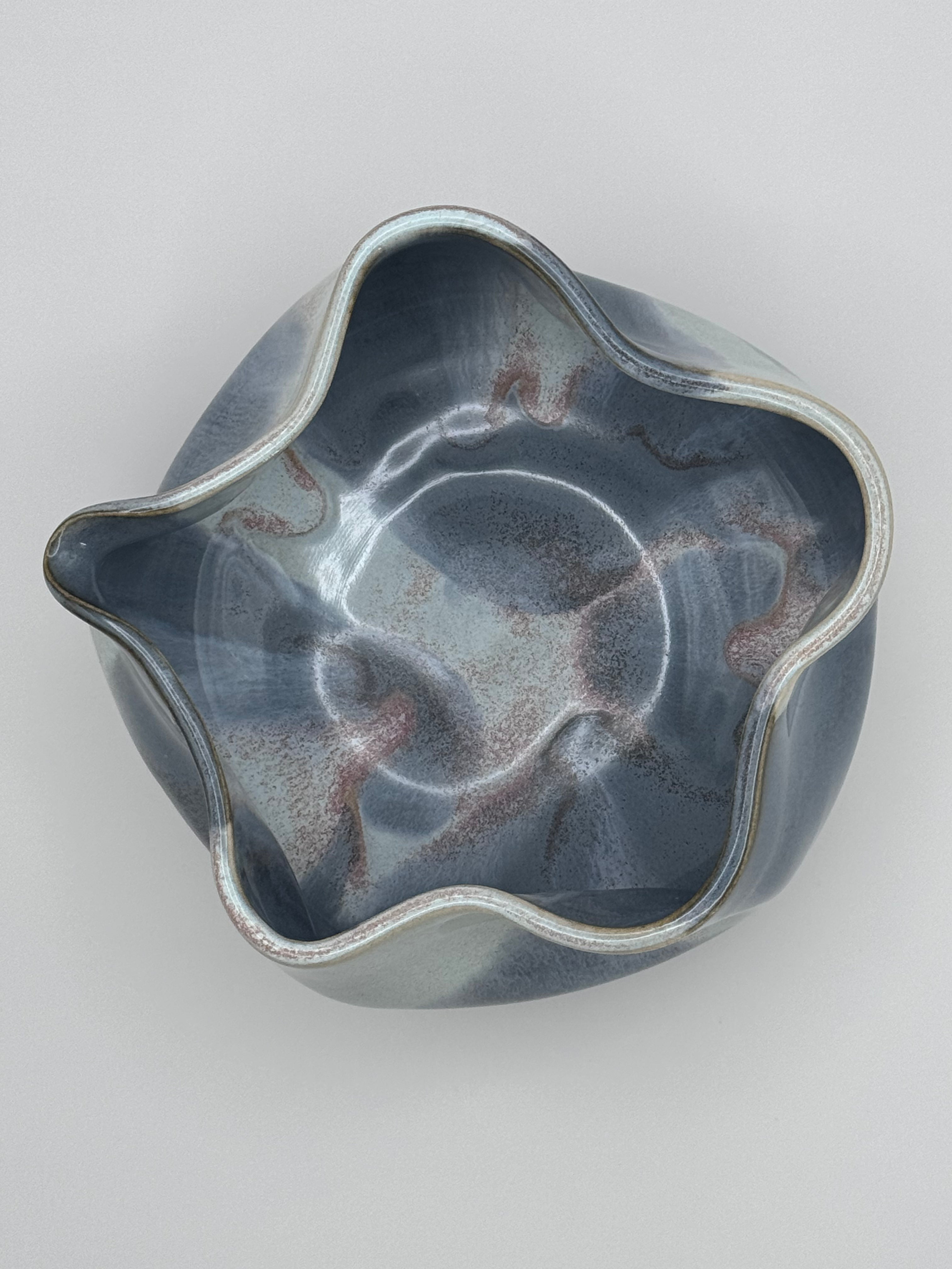 Top view of A handmade ceramic bowl with a wavy rim and a small footed base, featuring a glossy glaze with abstract slated blue and light blue brushstrokes over a white clay base and dusty rose specks on the interior, set against a plain light gray background.