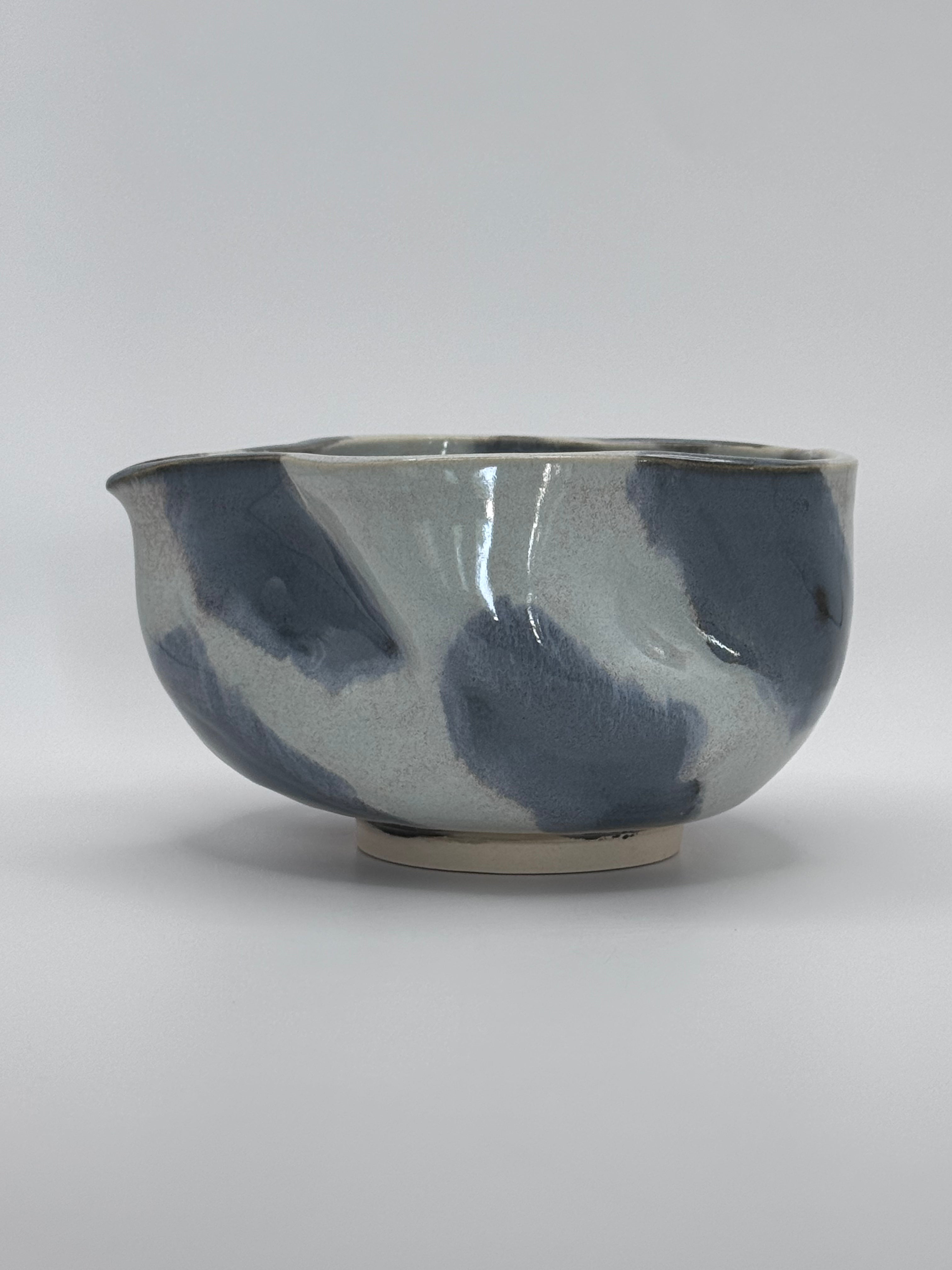 A handmade ceramic bowl with a wavy rim and a small footed base, featuring a glossy glaze with abstract slated blue and light blue brushstrokes over a white clay base, set against a plain light gray background.