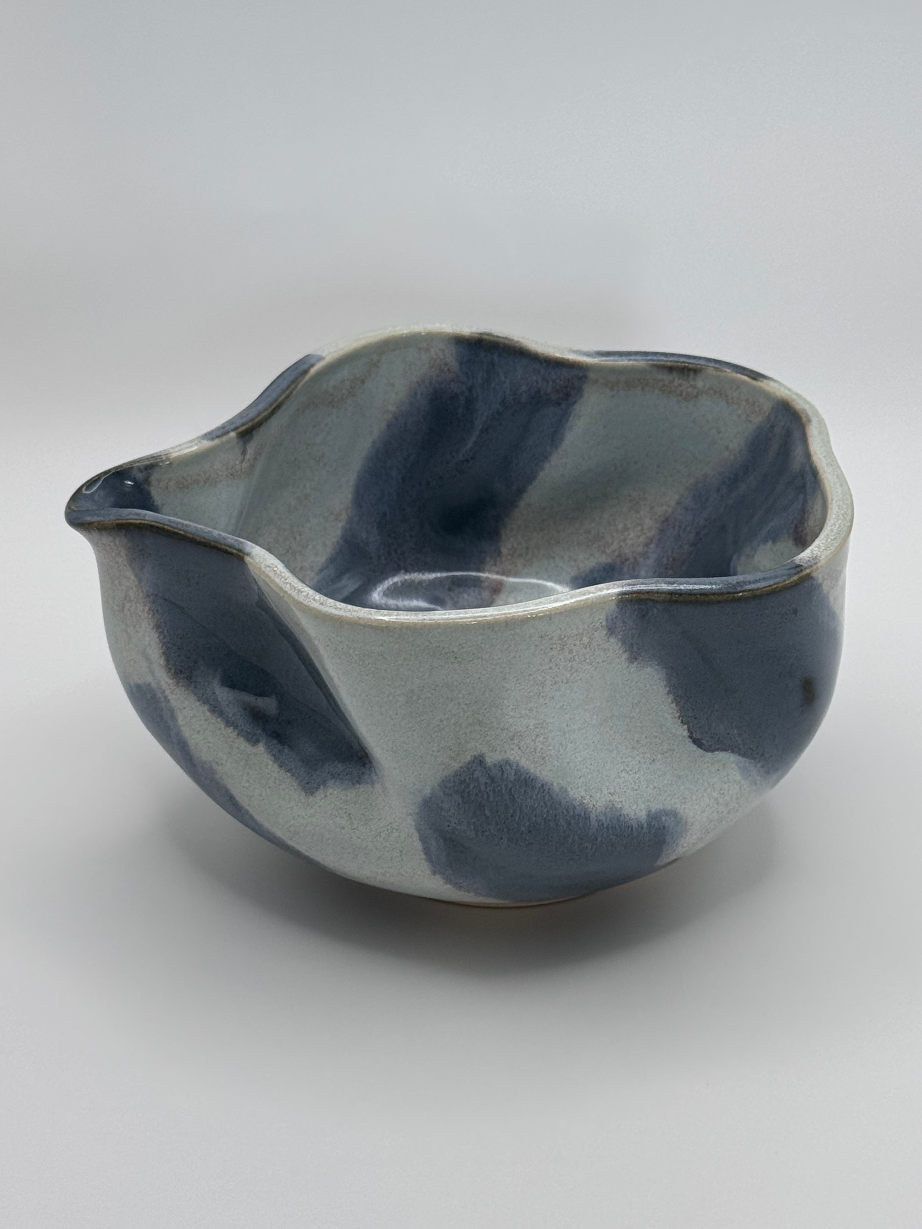 A handmade ceramic bowl with a wavy rim and a small footed base, featuring a glossy glaze with abstract slated blue and light blue brushstrokes over a white clay base, set against a plain light gray background.