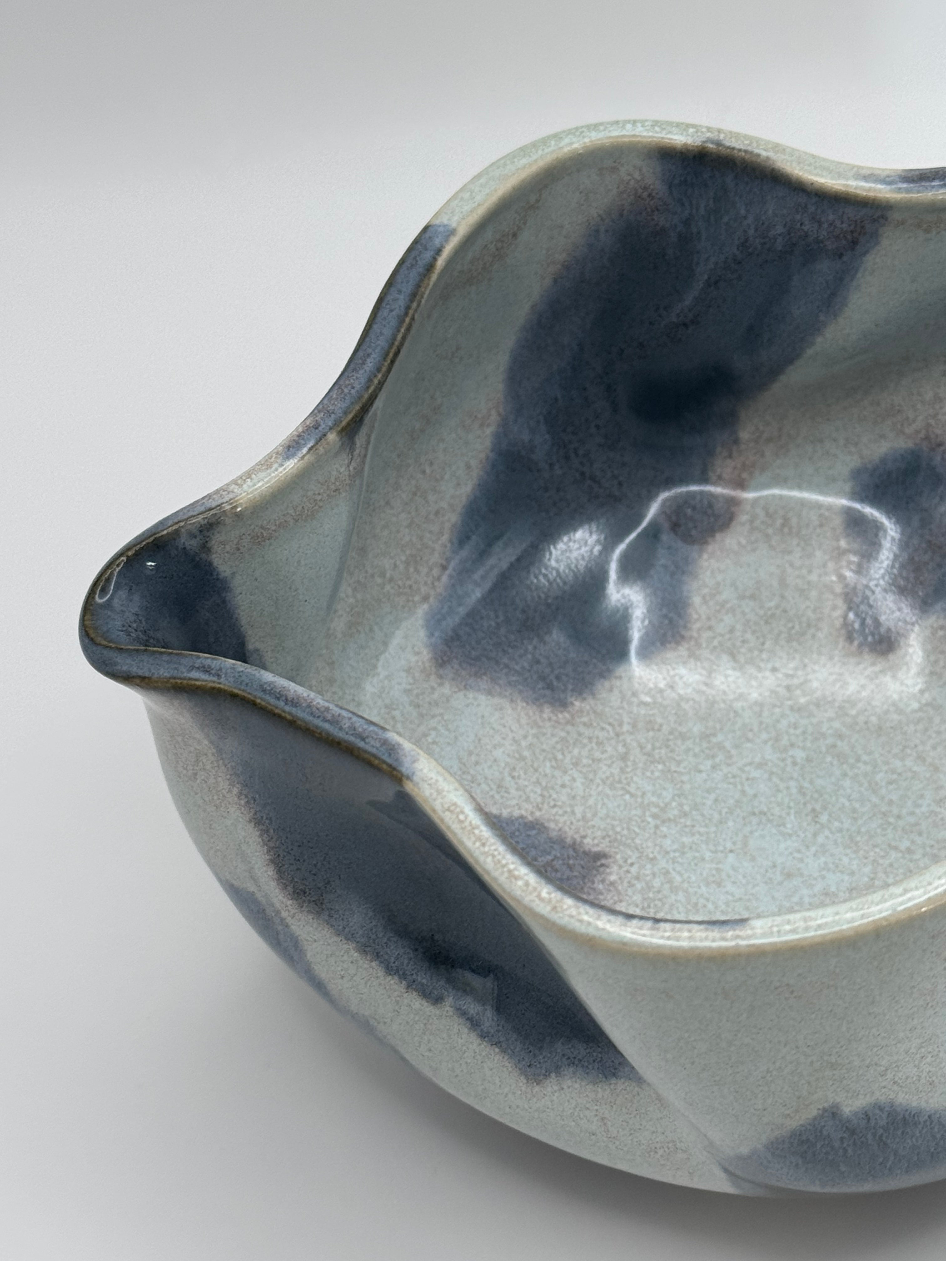A close-up of A handmade ceramic bowl with a wavy rim and a small footed base, featuring a glossy glaze with abstract slated blue and light blue brushstrokes over a white clay base, set against a plain light gray background.