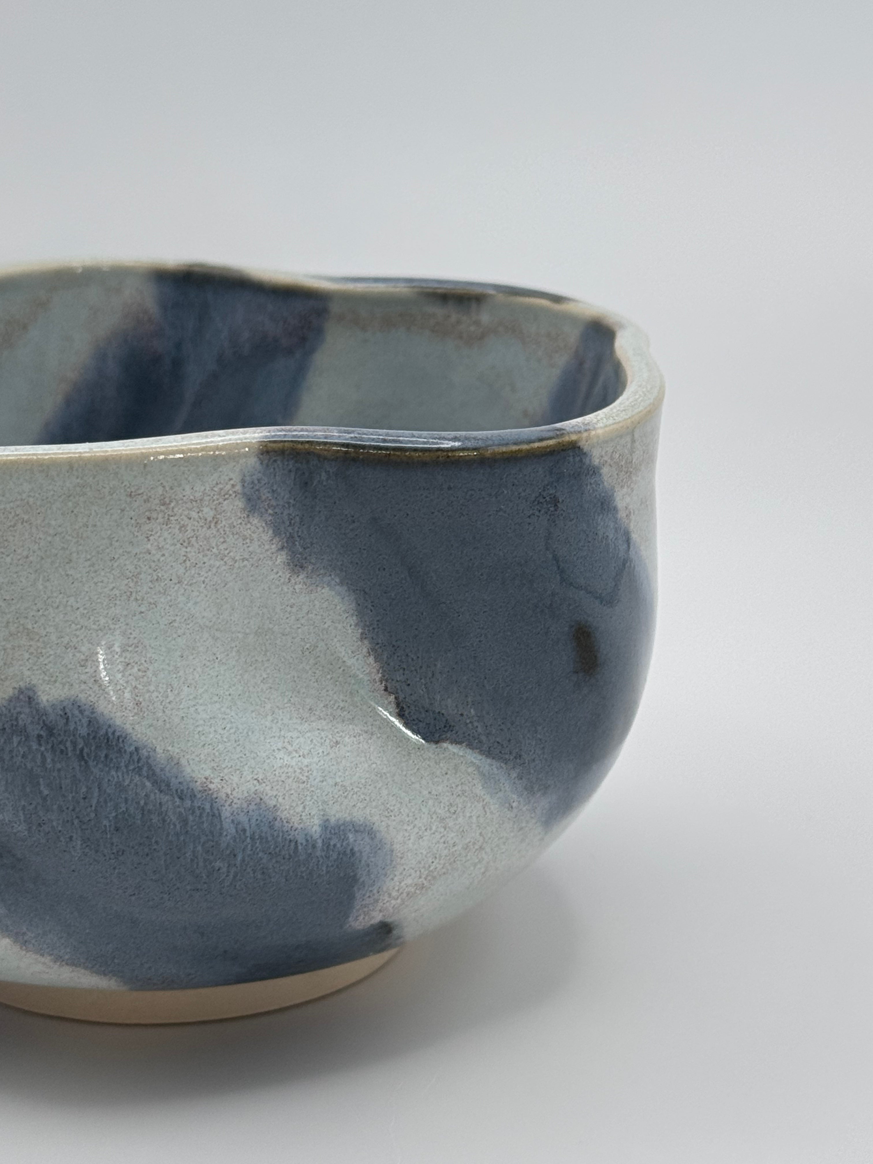 A close-up of A handmade ceramic bowl with a wavy rim and a small footed base, featuring a glossy glaze with abstract slated blue and light blue brushstrokes over a white clay base, set against a plain light gray background.