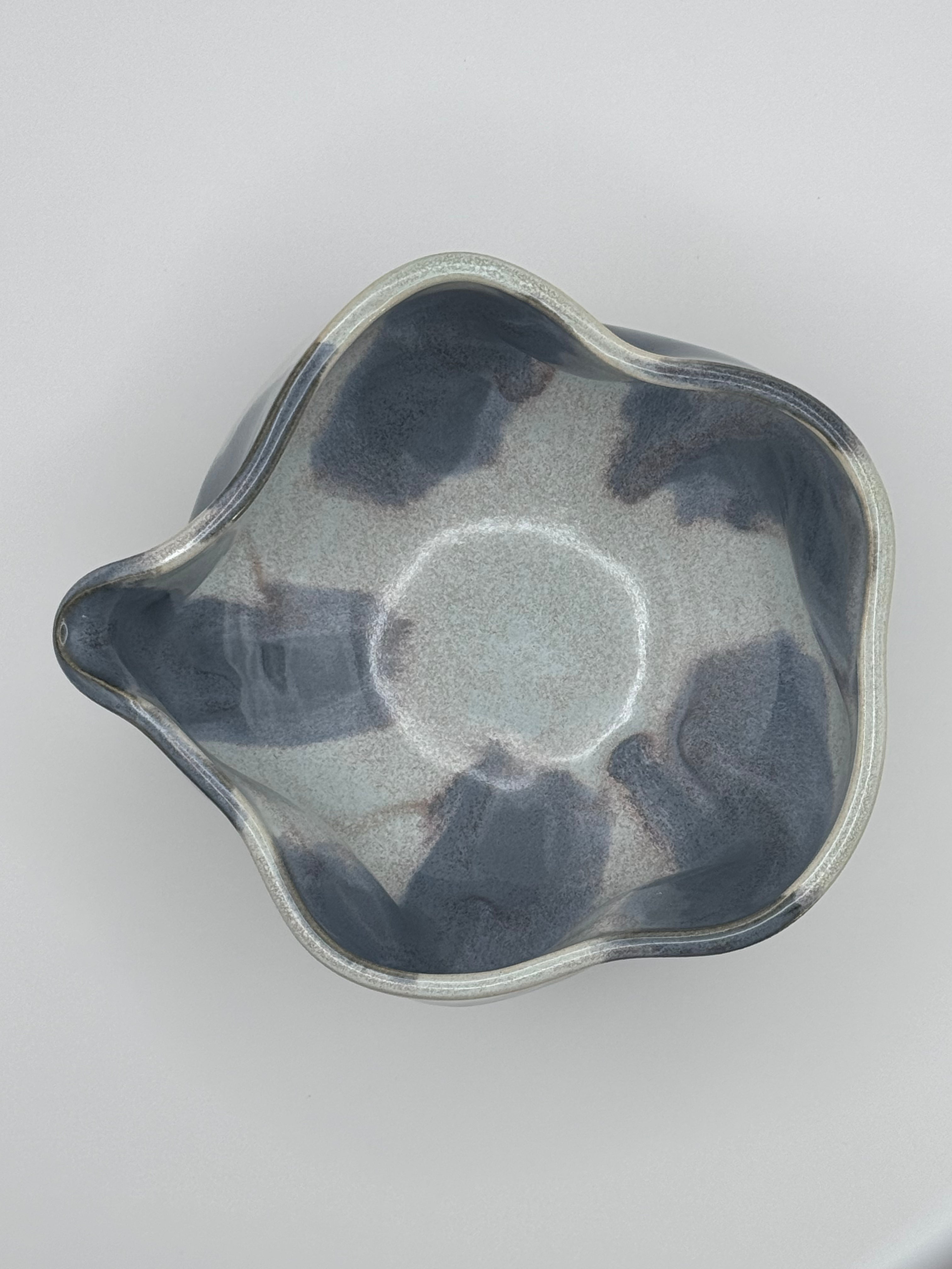 Top view of A handmade ceramic bowl with a wavy rim and a small footed base, featuring a glossy glaze with abstract slated blue and light blue brushstrokes over a white clay base and with dusty rose specks on the interior, set against a plain light gray background.