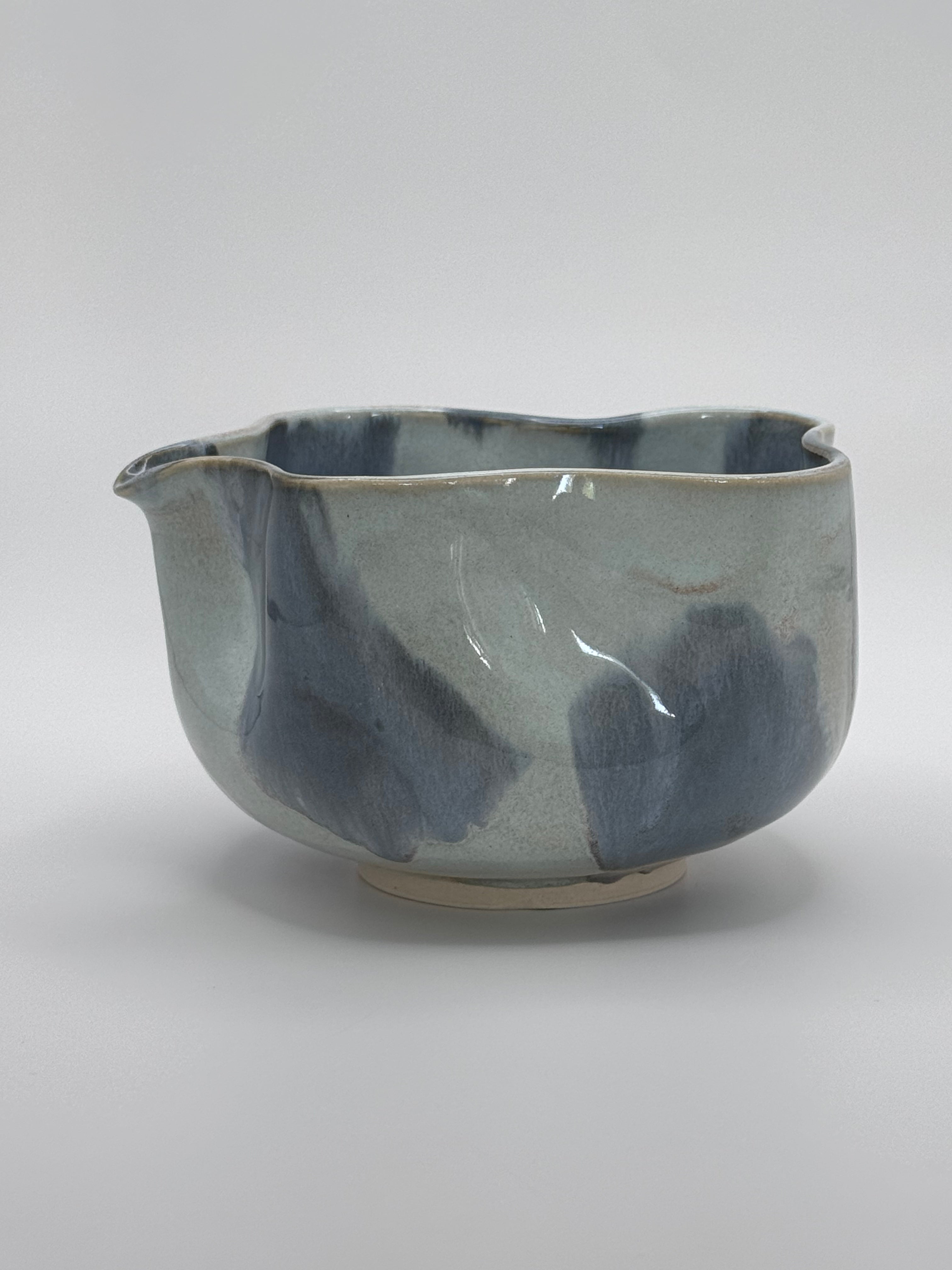A handmade ceramic bowl with a wavy rim and a small footed base, featuring a glossy glaze with abstract slated blue and light blue brushstrokes over a white clay base, set against a plain light gray background.