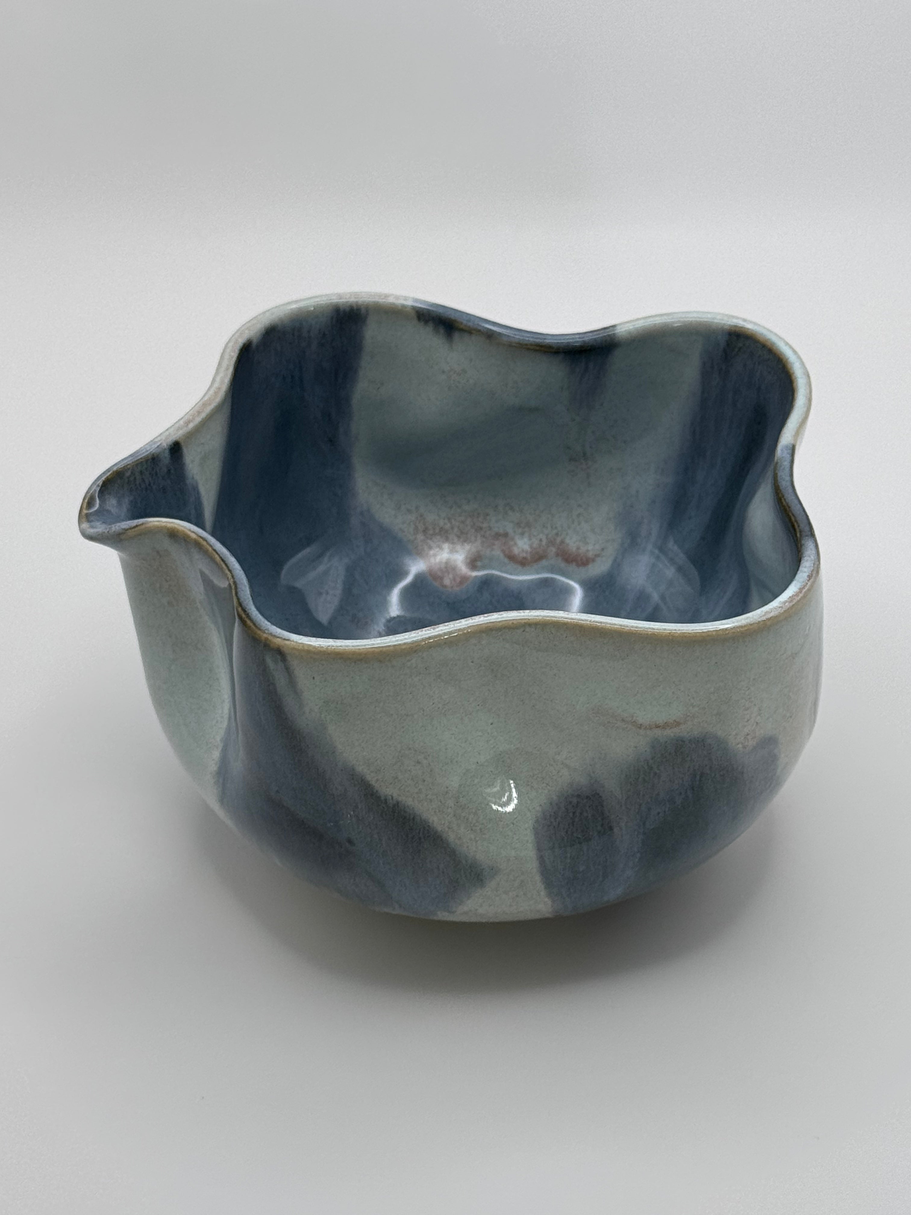A handmade ceramic bowl with a wavy rim and a small footed base, featuring a glossy glaze with abstract slated blue and light blue brushstrokes over a white clay base, set against a plain light gray background.