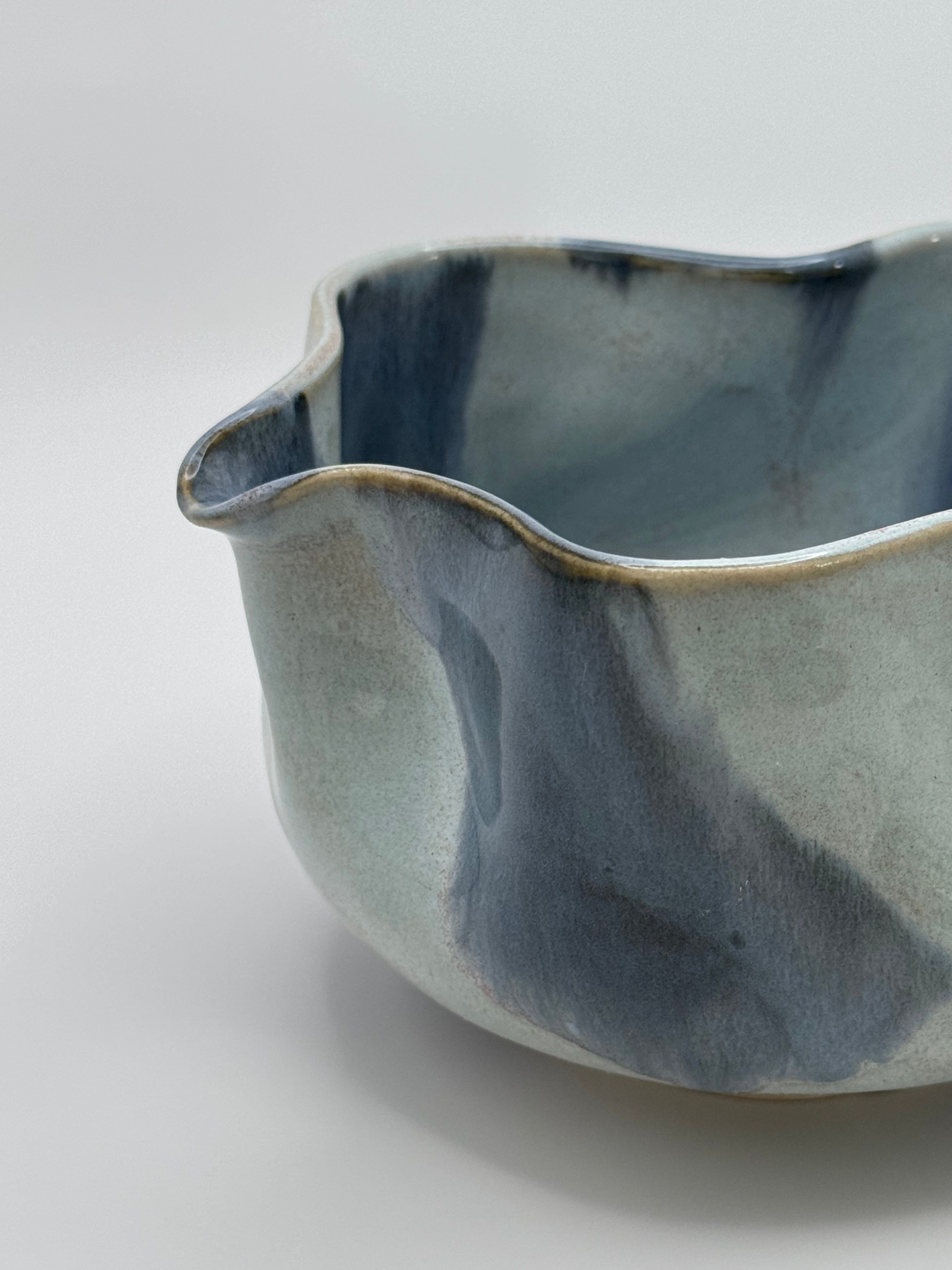 A close-up of A handmade ceramic bowl with a wavy rim and a small footed base, featuring a glossy glaze with abstract slated blue and light blue brushstrokes over a white clay base, set against a plain light gray background.