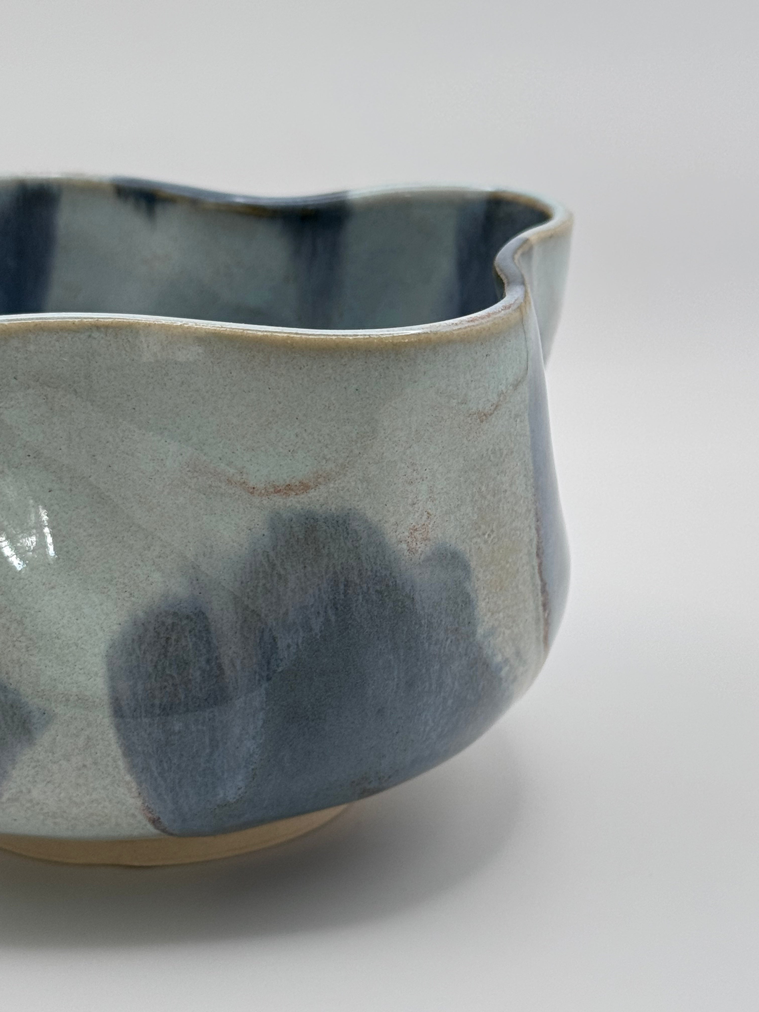 A close-up of A handmade ceramic bowl with a wavy rim and a small footed base, featuring a glossy glaze with abstract slated blue and light blue brushstrokes over a white clay base, set against a plain light gray background.