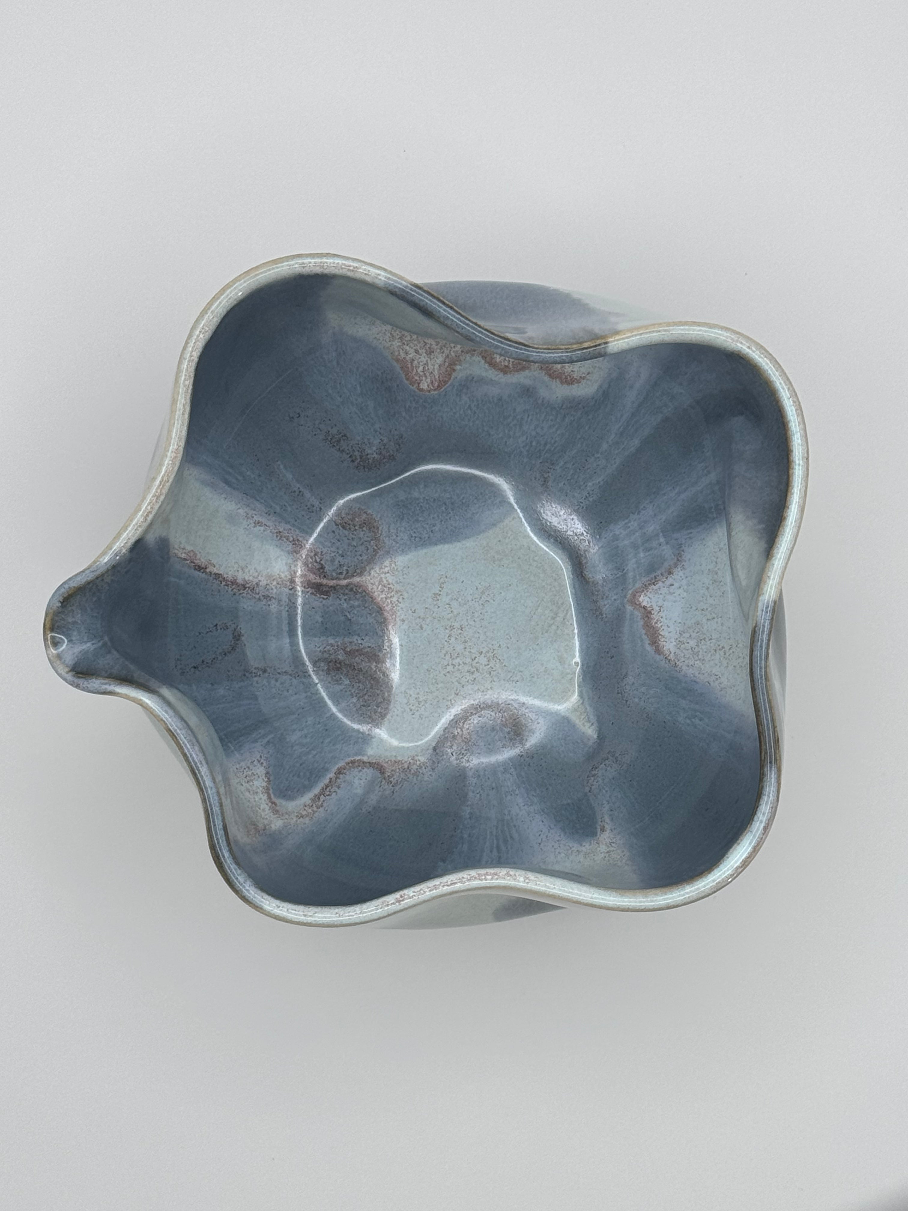 top view of A handmade ceramic bowl with a wavy rim and a small footed base, featuring a glossy glaze with abstract slated blue and light blue brushstrokes over a white clay base and with dusty rose specks on the interior, set against a plain light gray background.