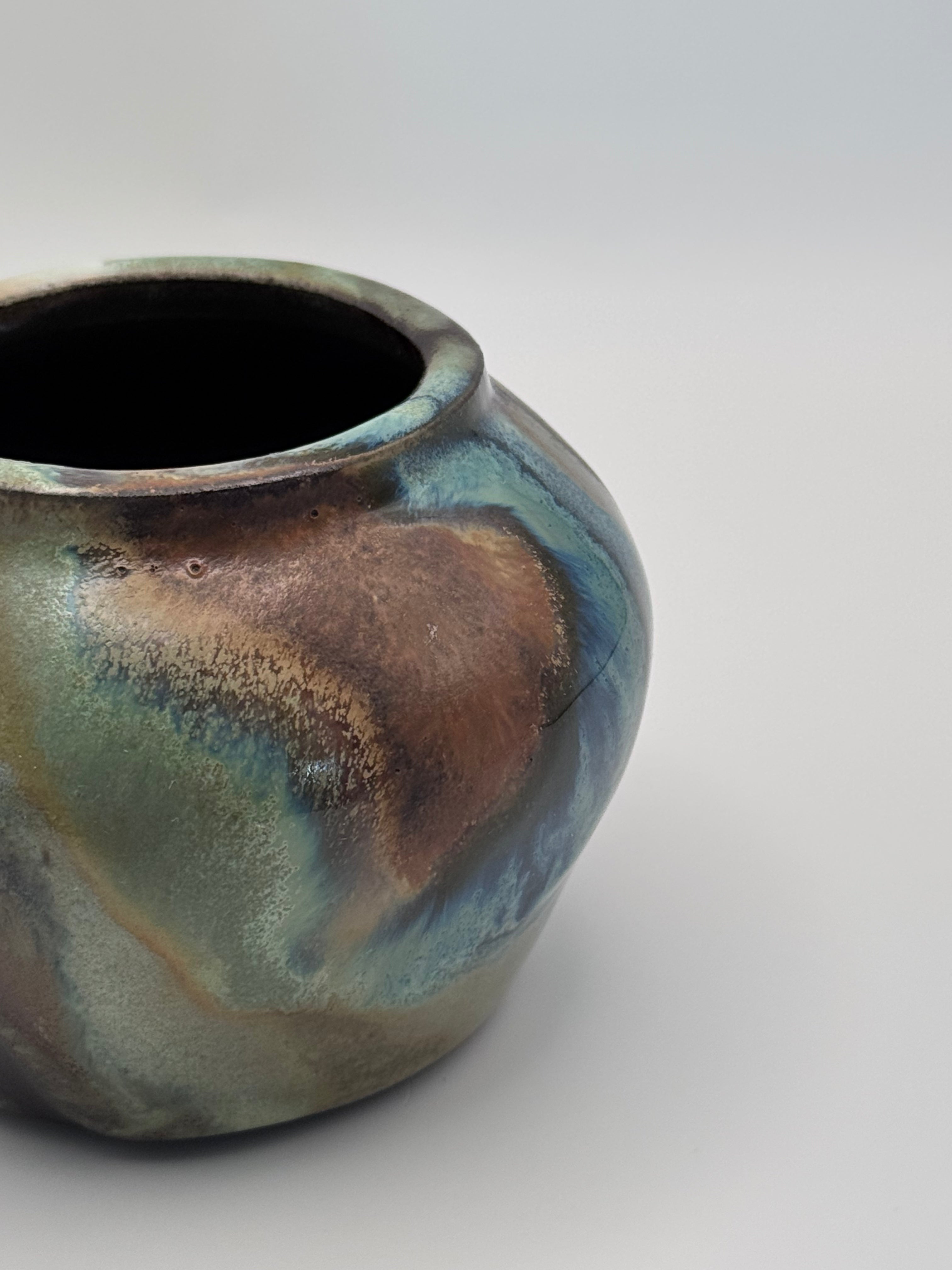 Close up view of small ceramic bud vase with rounded shoulders, marbled dark brown, white, and green celadon glaze with metallic crystalline accents.