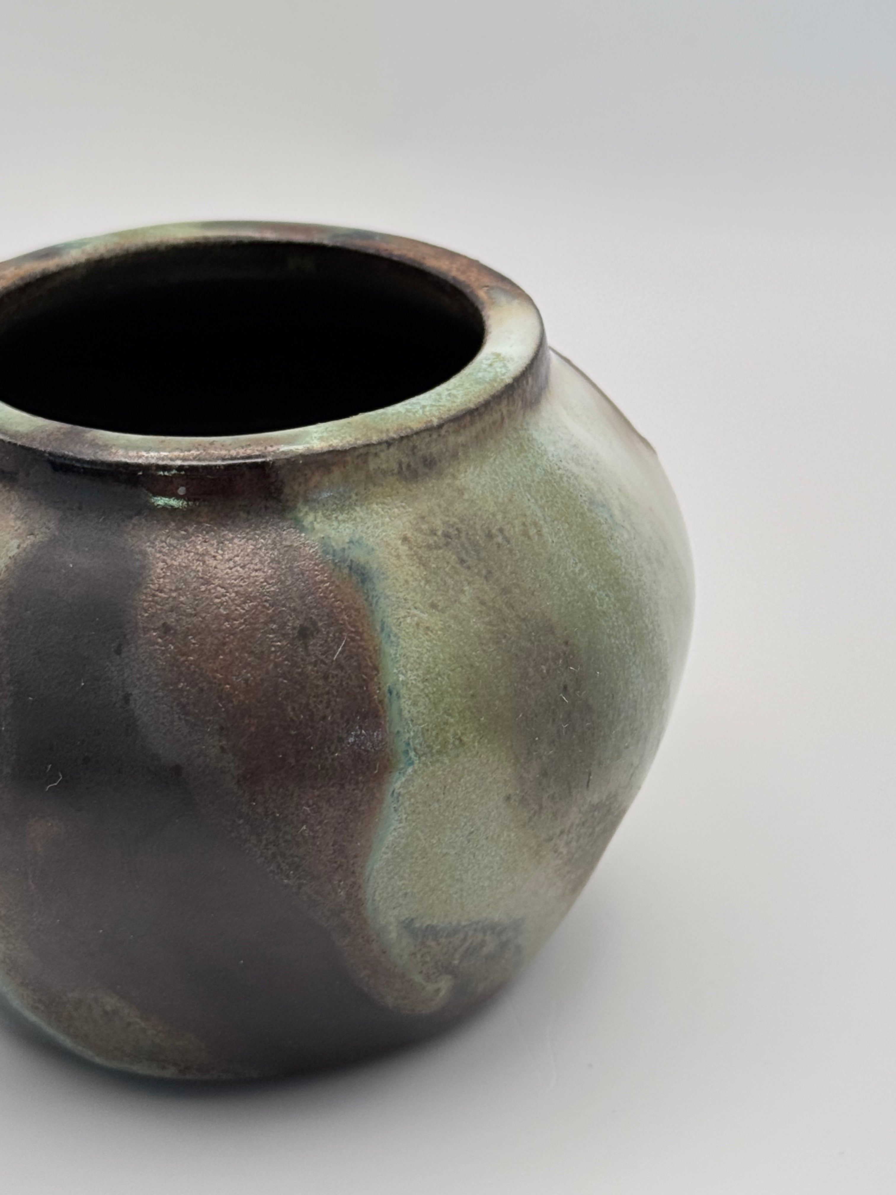 Close up view of small ceramic bud vase with rounded shoulders, marbled dark brown, white, and green celadon glaze with metallic crystalline accents.