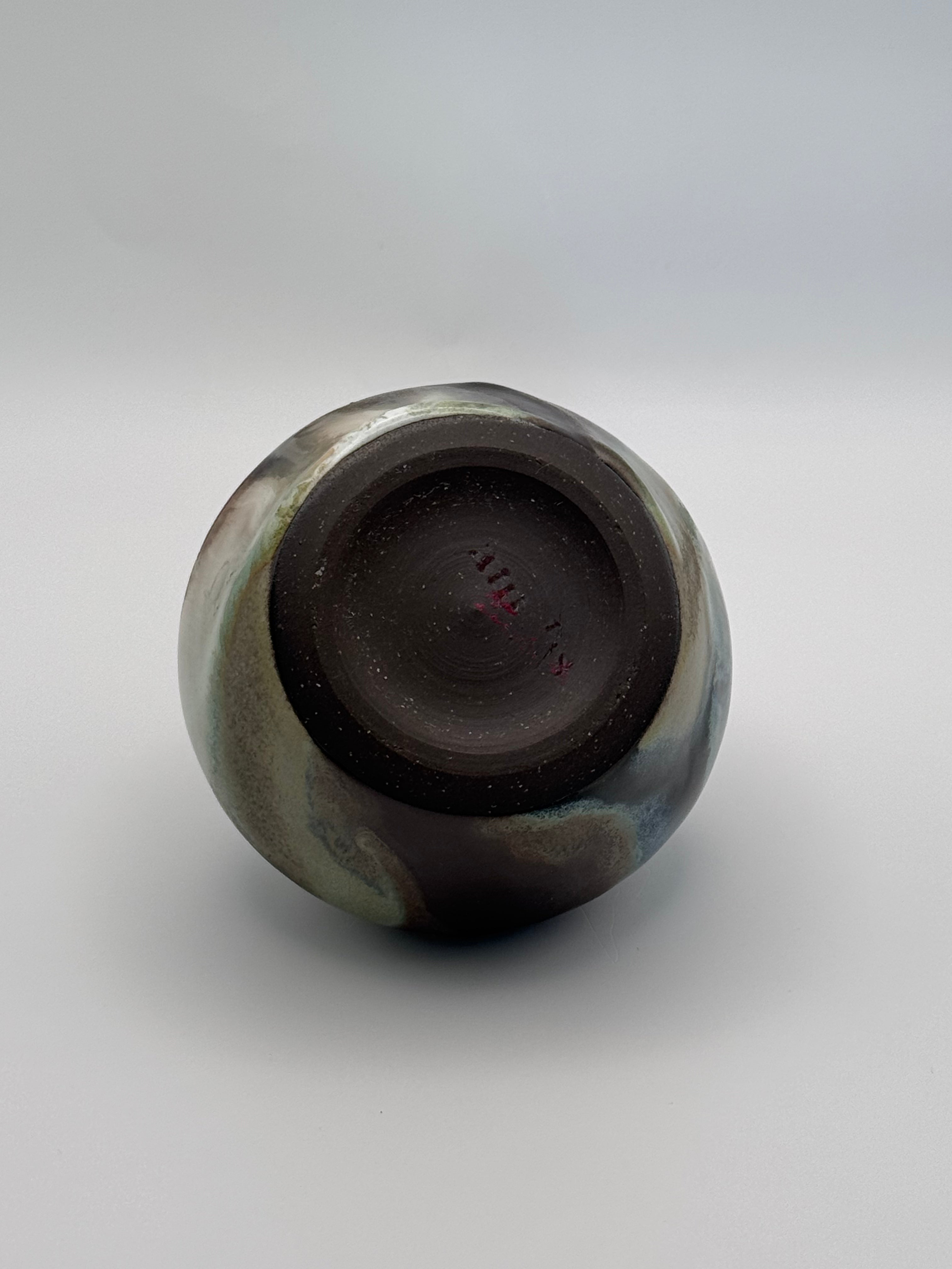 View of foot of small ceramic bud vase with rounded shoulders, marbled dark brown, white, and green celadon glaze with metallic crystalline accents.