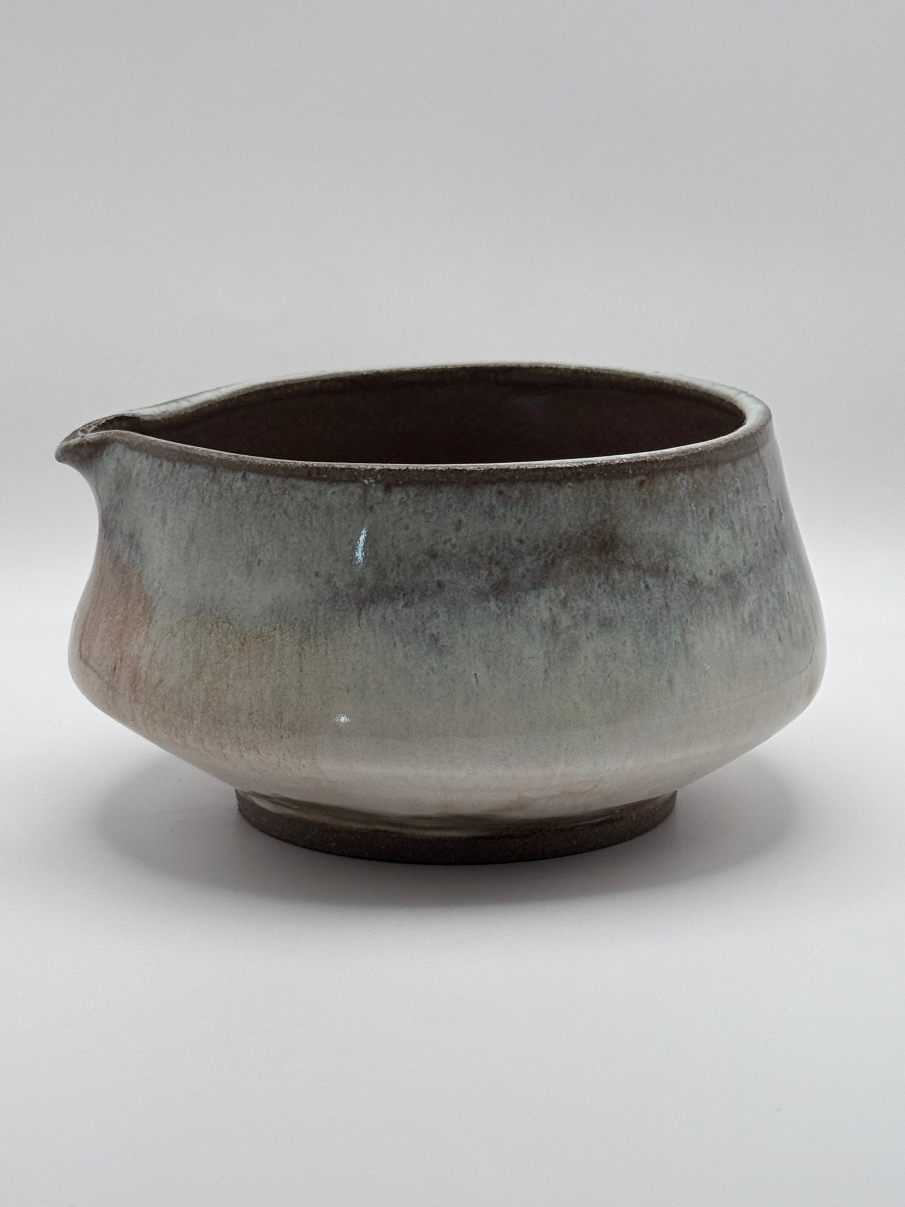 A handmade ceramic pitcher with a small pouring spout, featuring a rustic glaze that transitions from a dark brown interior to a mottled gray and beige exterior, set against a neutral gray background.
