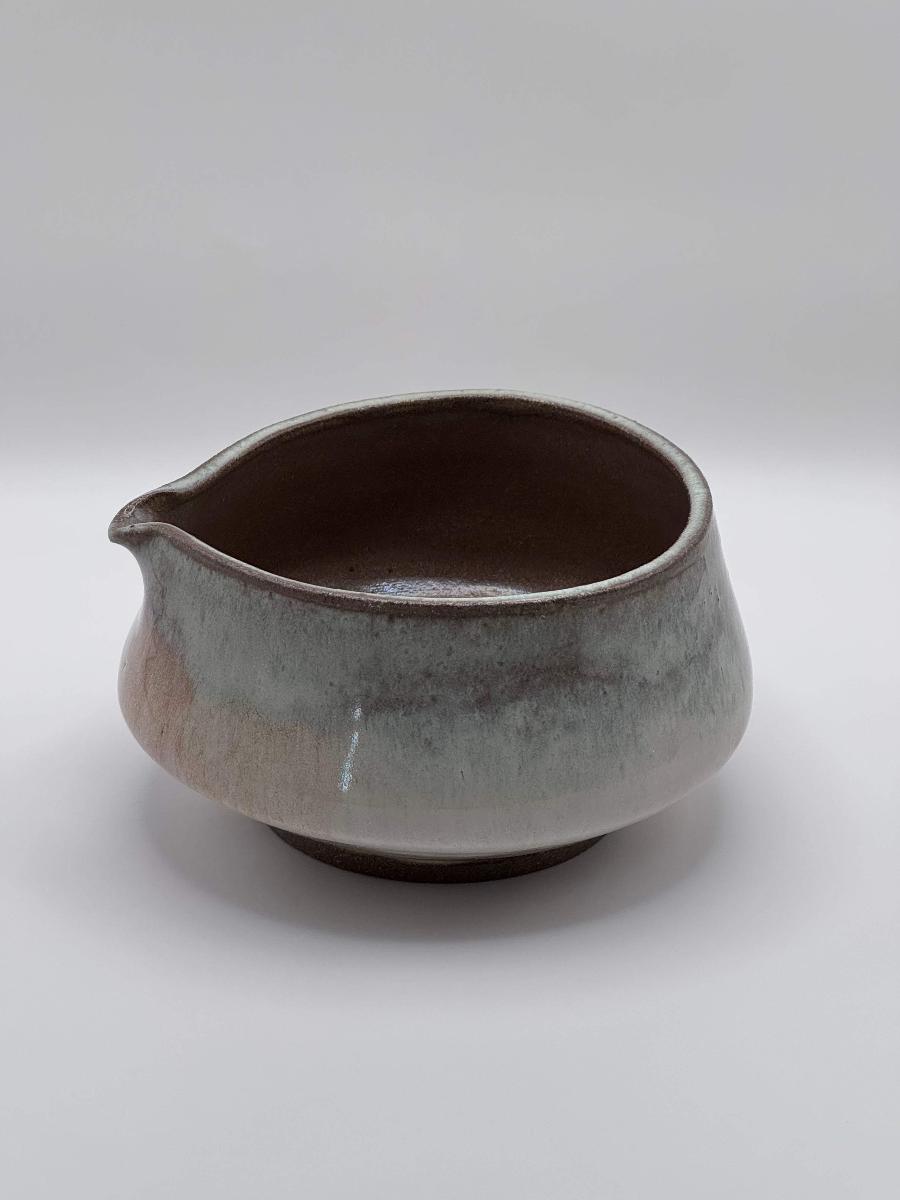 A handmade ceramic pitcher with a small pouring spout, featuring a rustic glaze that transitions from a warm brown interior to a textured gray and beige exterior, set against a neutral gray background.