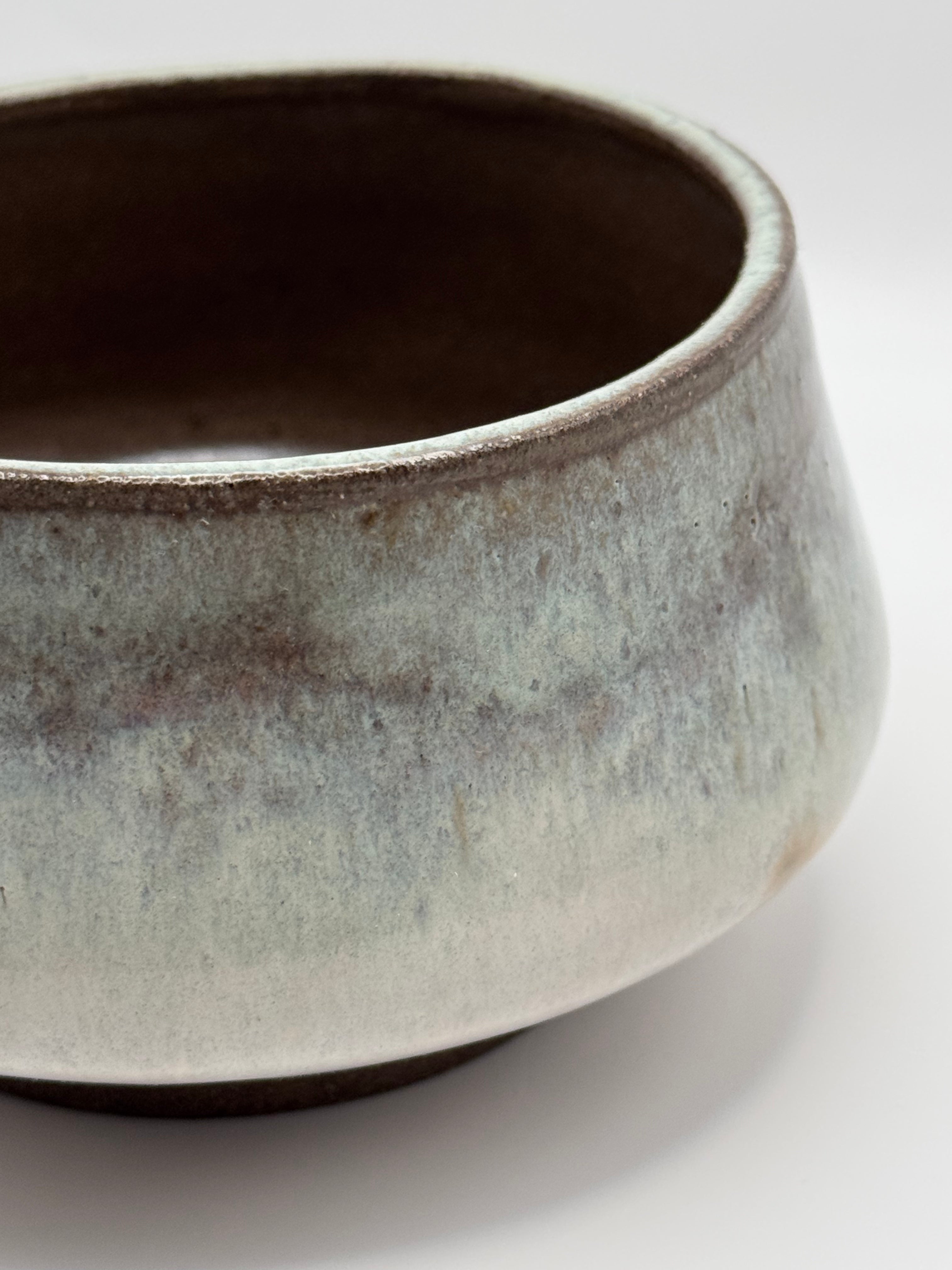 A close-up of a handmade ceramic bowl with a textured, rustic glaze in muted gray, brown, and off-white tones, set against a plain white background.