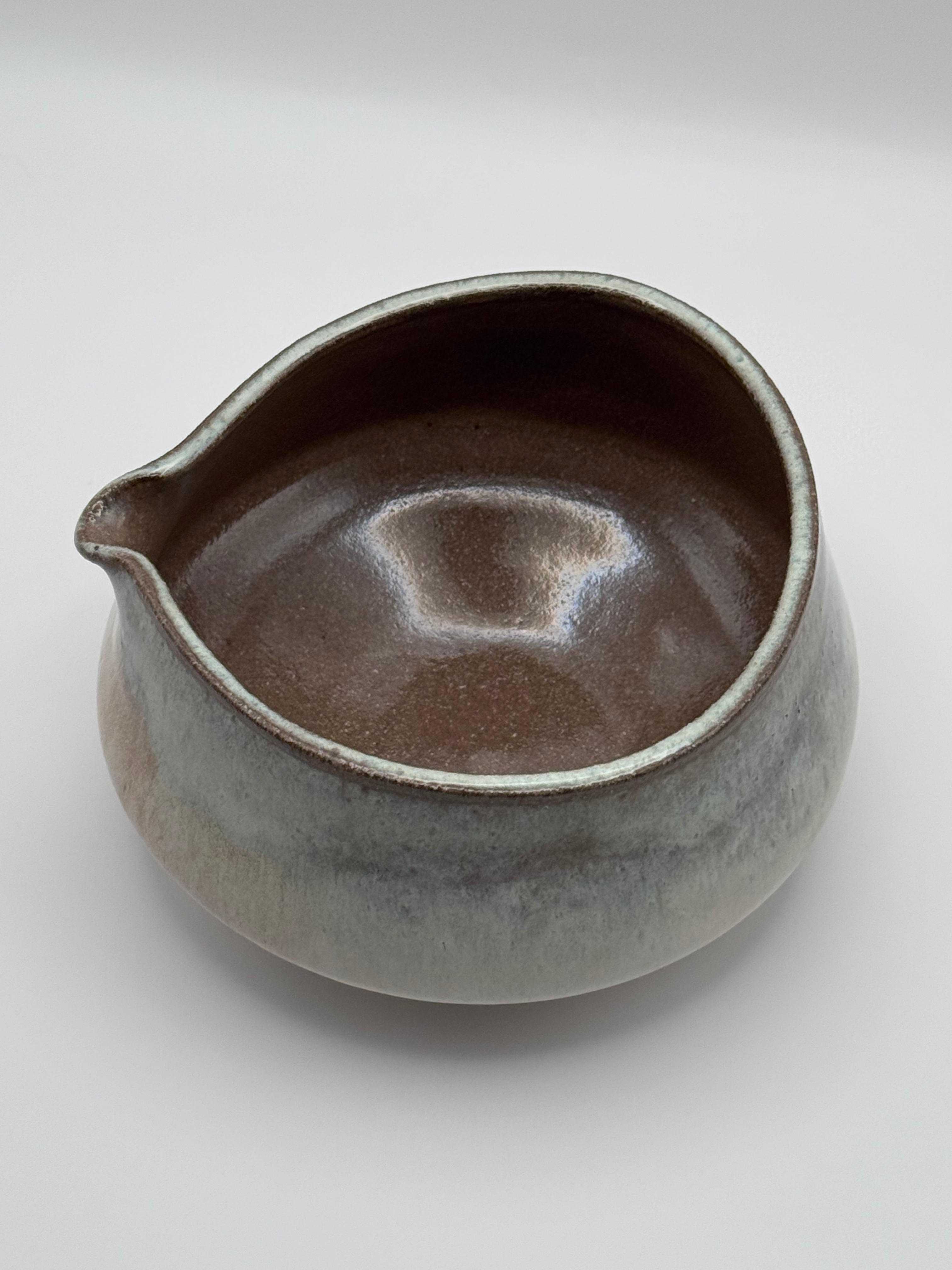 A handmade ceramic pitcher with a small pouring spout, featuring a rustic glaze that transitions from a dark brown interior to a speckled gray and beige exterior, set against a plain light gray background.