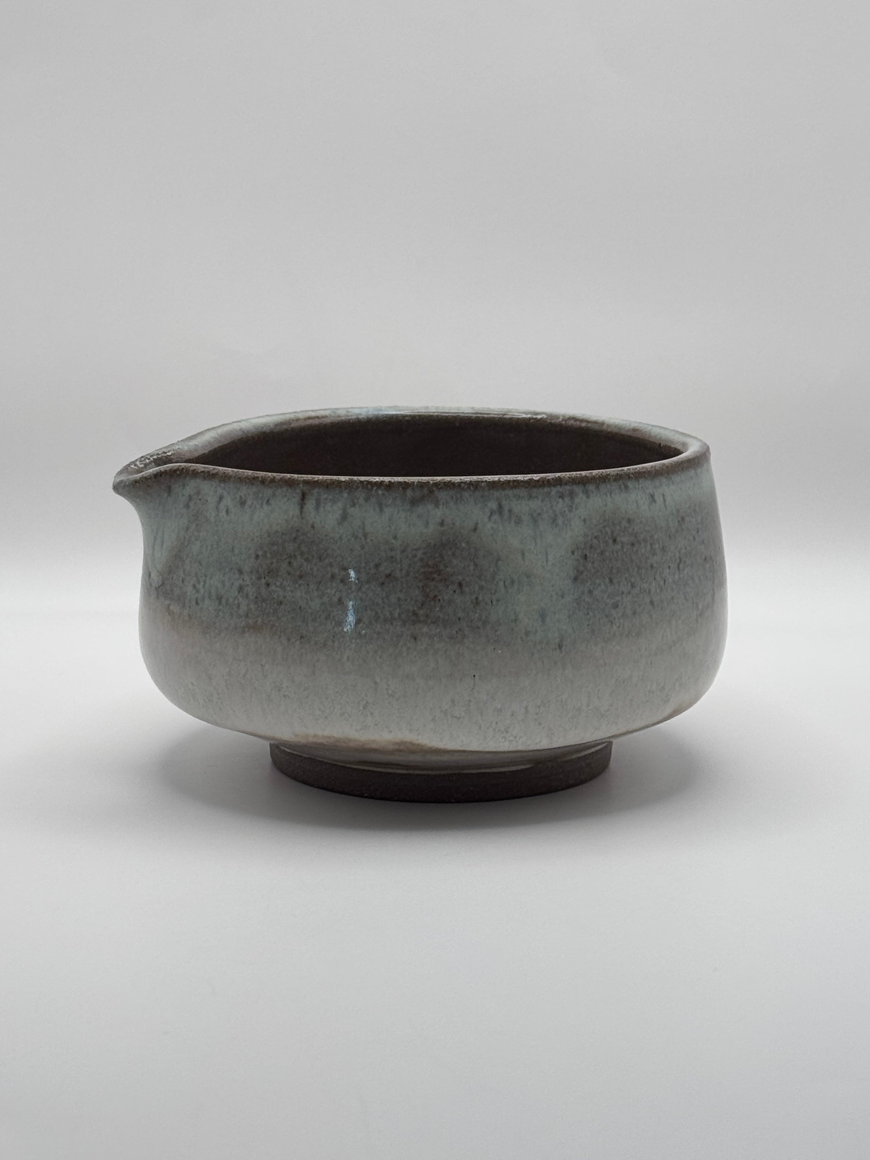 A handmade ceramic bowl with a small pouring spout, featuring a speckled gray and blue-green glaze with a dark brown interior and base, set against a neutral gray background.