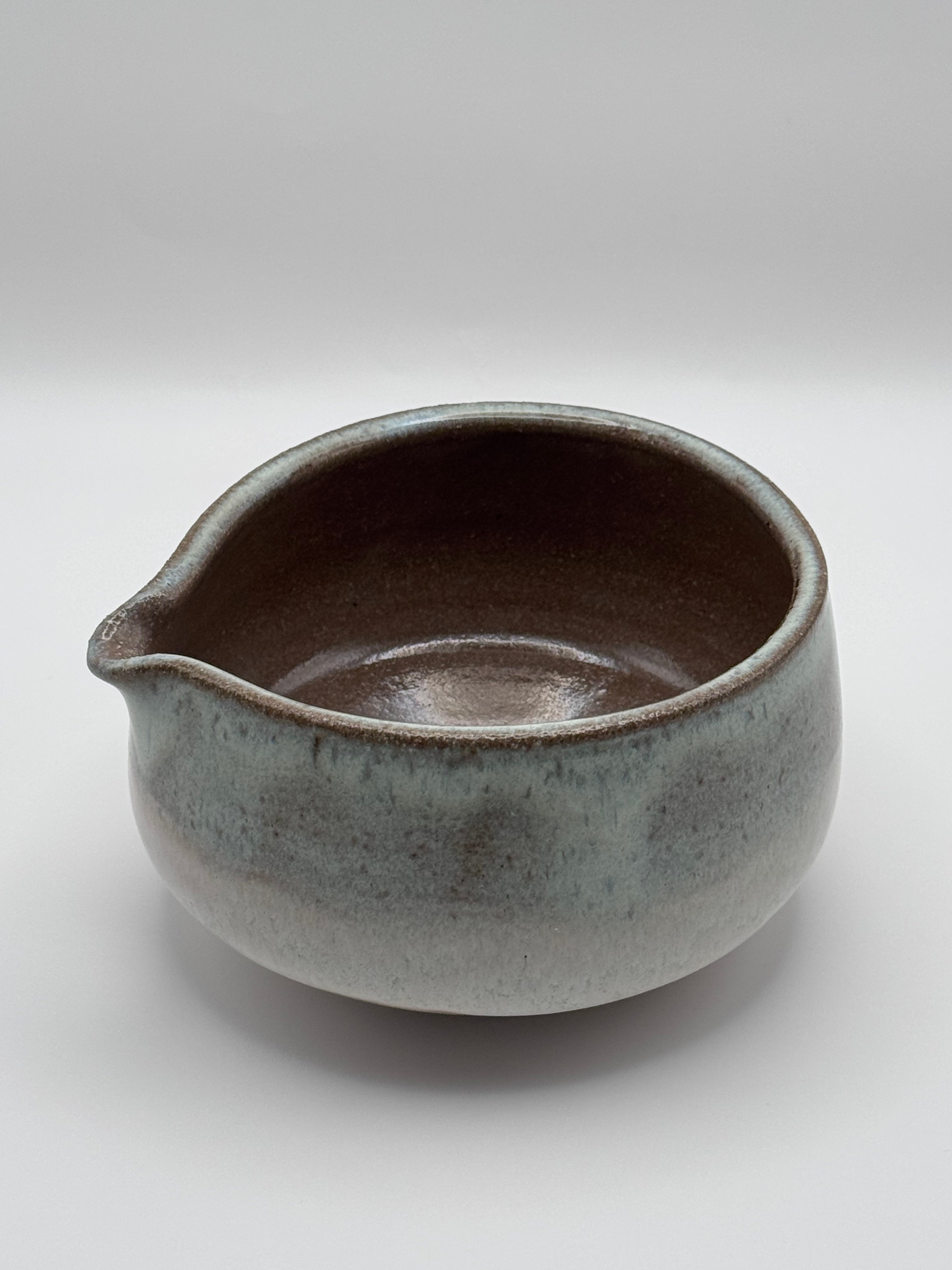 A small ceramic pitcher with a rounded body, a small pouring spout on the left, and a glossy speckled glaze transitioning from pale blue-gray to dark brown, photographed on a light gray background.