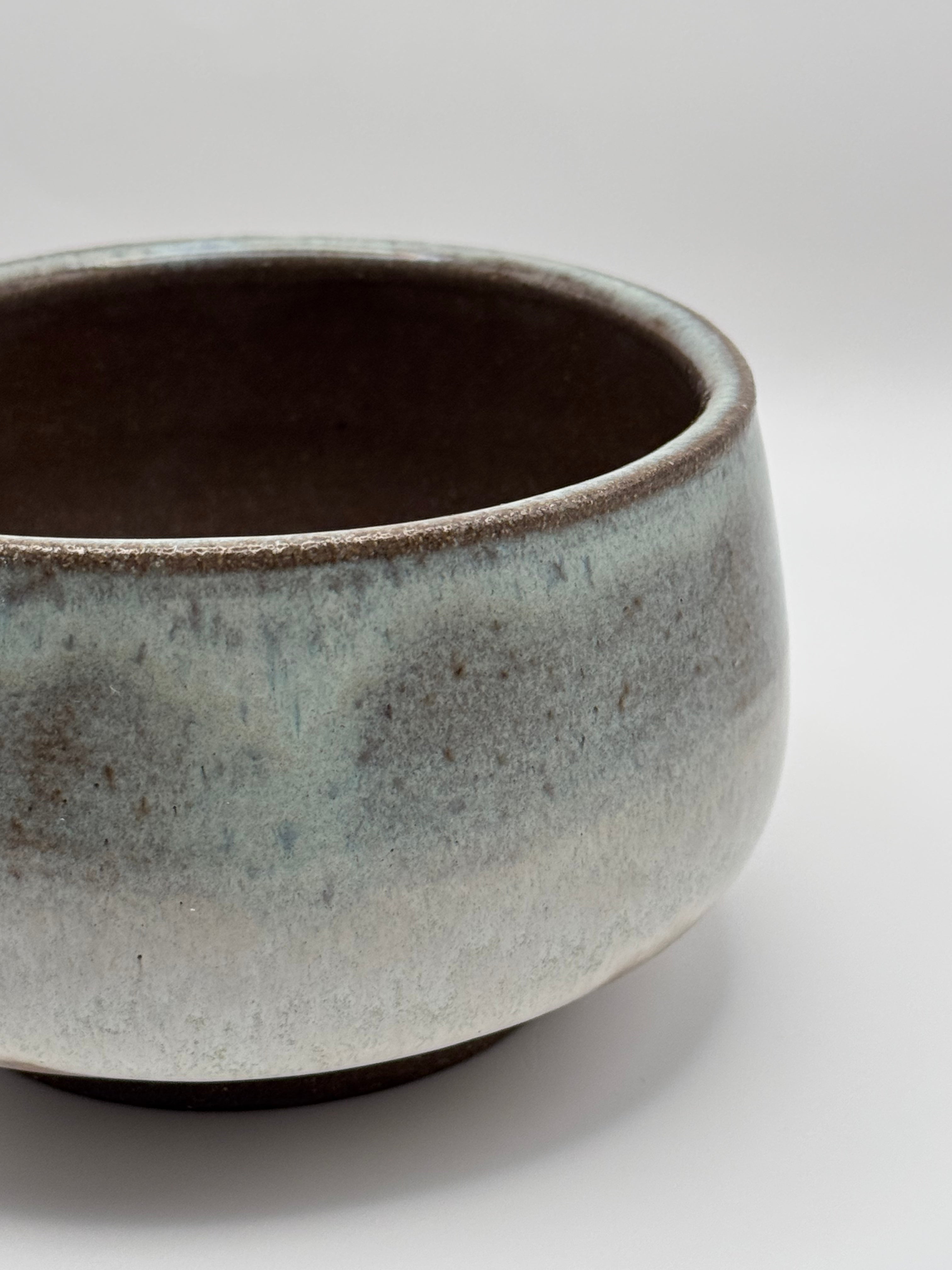 A close-up of a handmade ceramic bowl with a textured, speckled glaze transitioning from a light blue-gray exterior to a dark brown interior, set against a neutral light gray background.