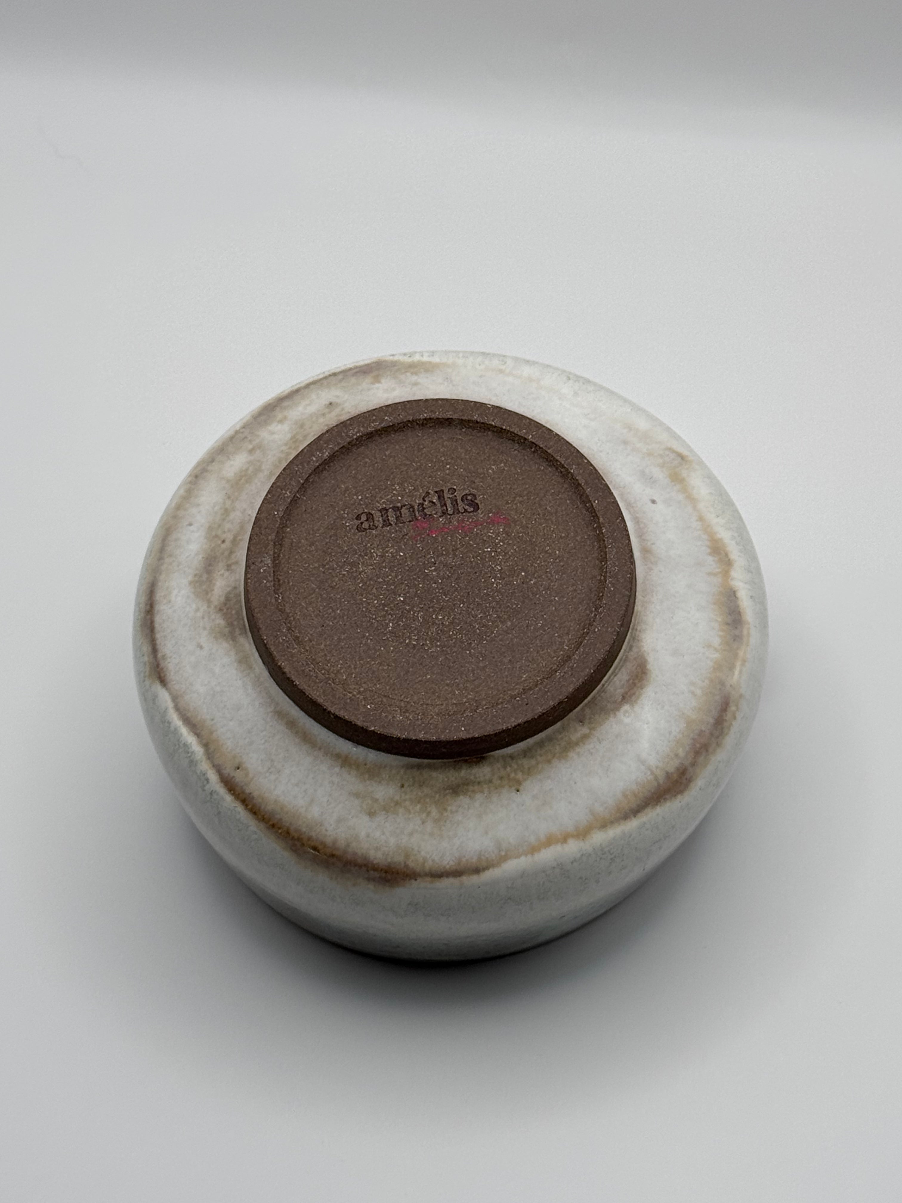 The bottom view of a handmade ceramic bowl with a textured, rustic finish. The exterior features a mottled glaze of off-white and earthy brown, while the center displays a circular brown base stamped with the brand name 'amelis'.