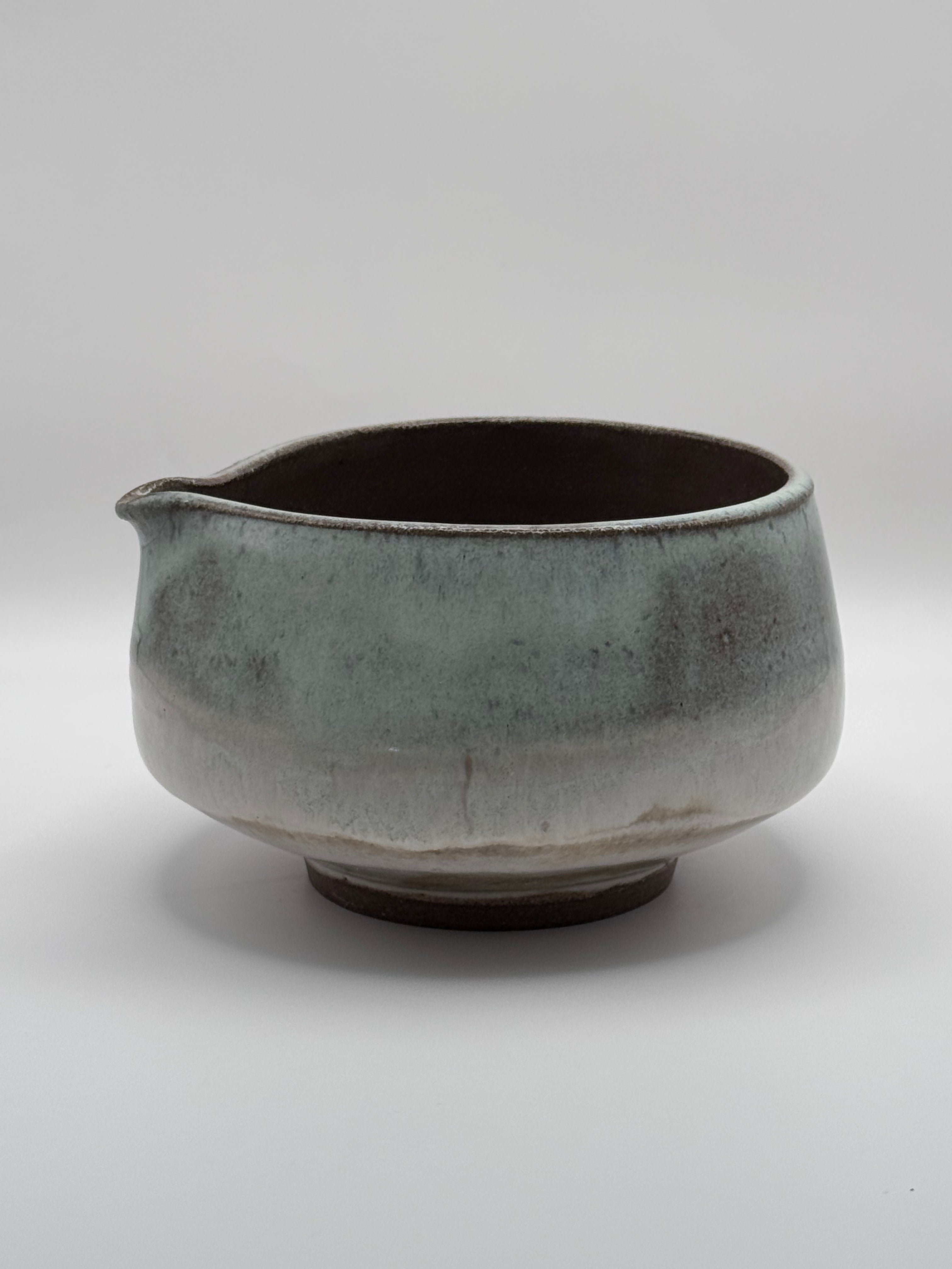 A handmade ceramic pitcher with a small pouring spout, featuring a textured, mottled glaze in shades of gray, blue-green, and brown, set against a plain light gray background.