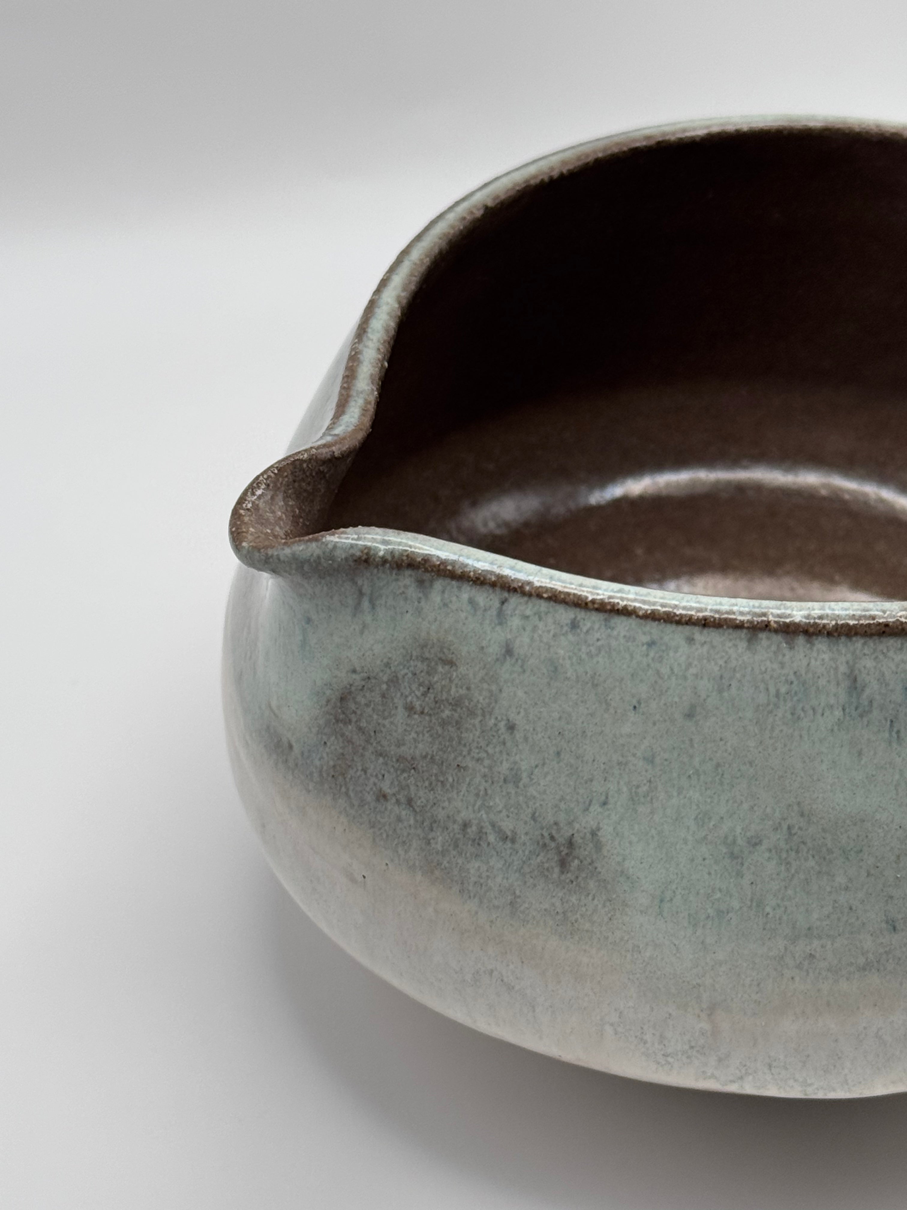 A close-up of a handmade ceramic pitcher with a textured, mottled glaze in shades of sage green, gray, and brown, set against a clean white background.