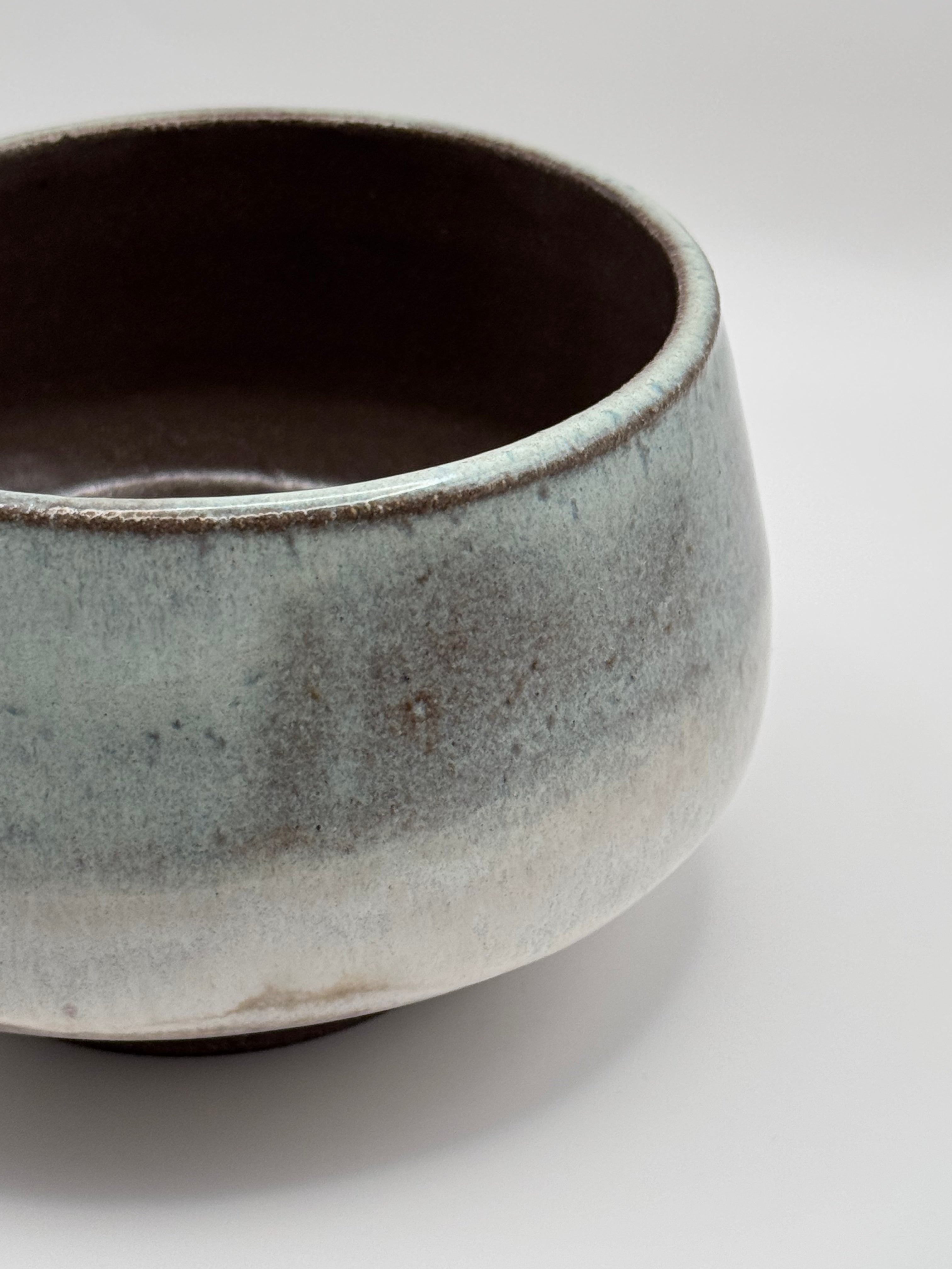 A close-up of a ceramic bowl with a textured, mottled glaze transitioning from pale blue-green to warm beige and brown, set against a clean white background.