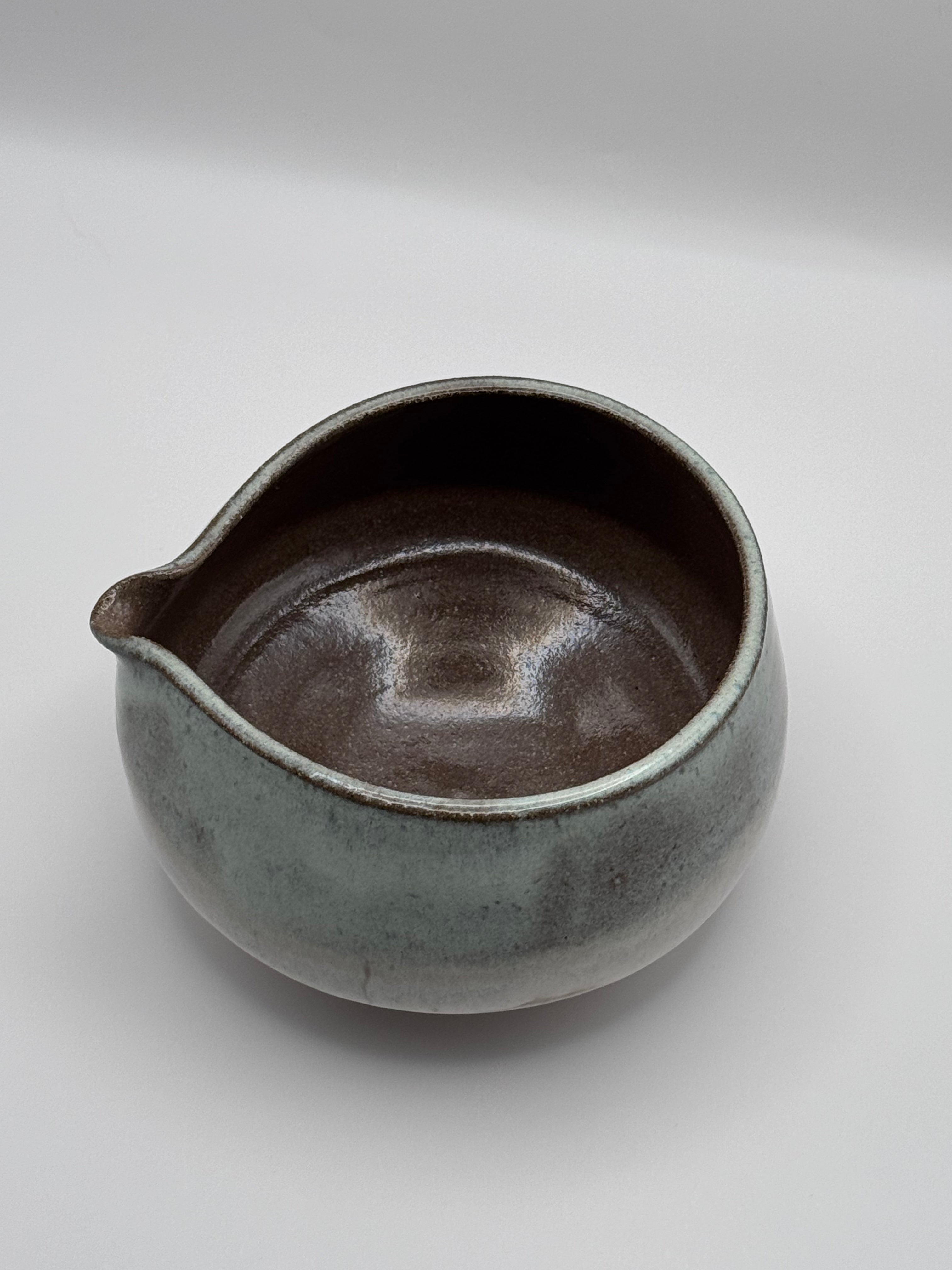 A handmade ceramic pitcher with a rounded body, a small pouring spout on the left, and a glossy glaze that transitions from a dark brown interior to a textured gray-green exterior, shown on a neutral light gray background.