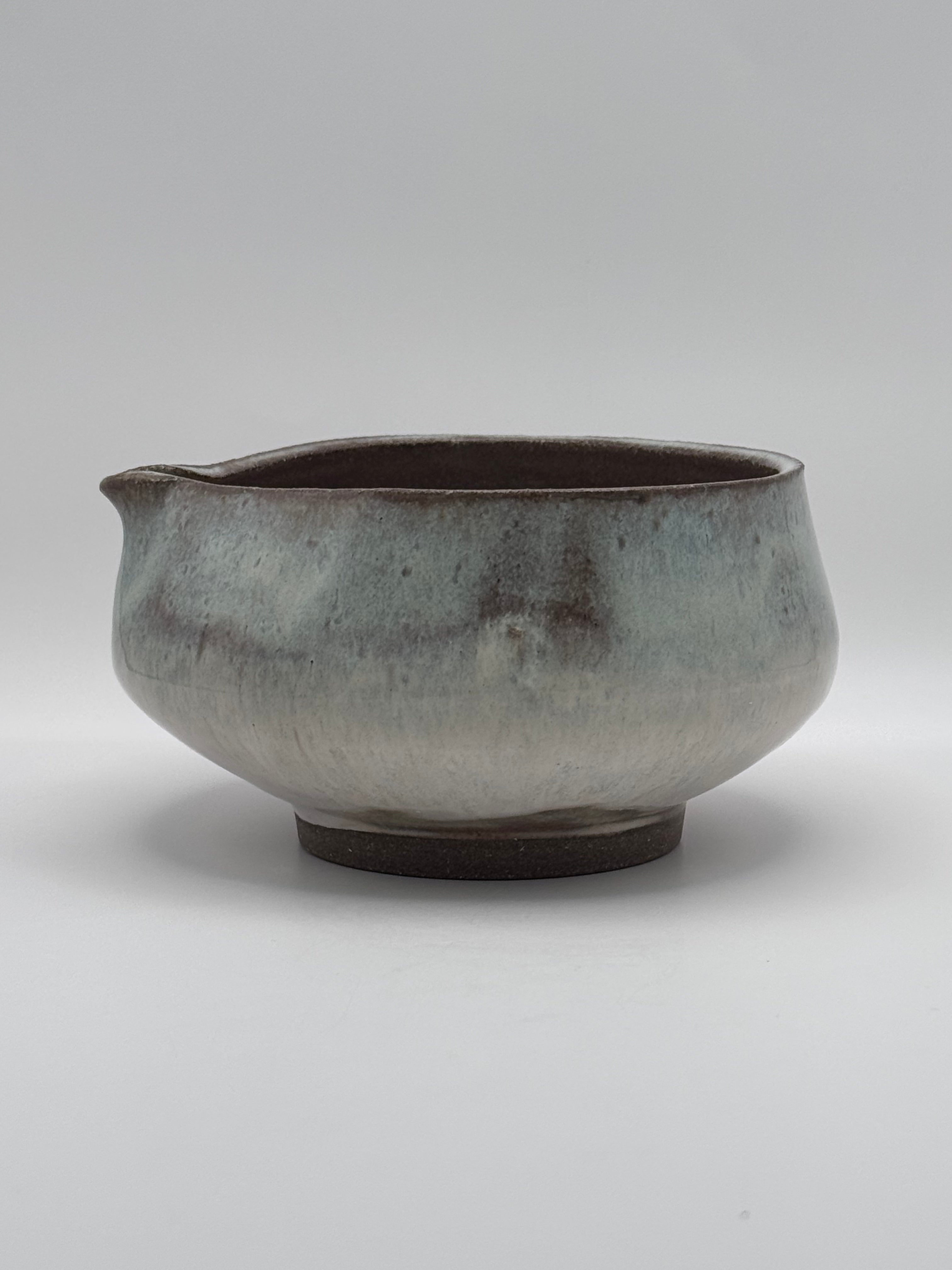A handmade ceramic pitcher with a small pouring spout, featuring a rustic glaze that transitions from a dark brown interior to a mottled gray and beige exterior, set against a neutral gray background.